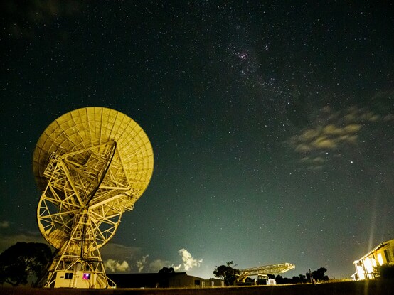 The night sky and a radio telescope and surrounding buildings.