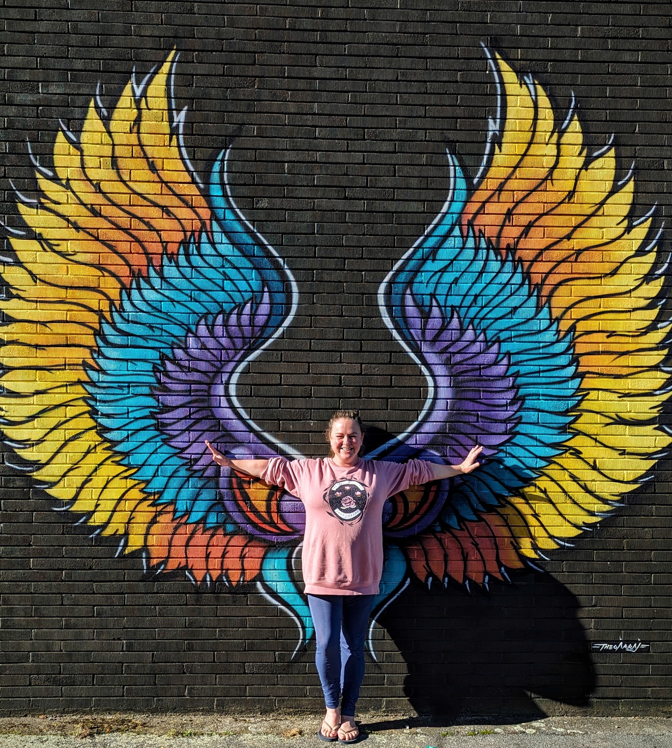 Large colourful 'angel wings' mural with a woman standing against it, arms out, as if the wings are hers.