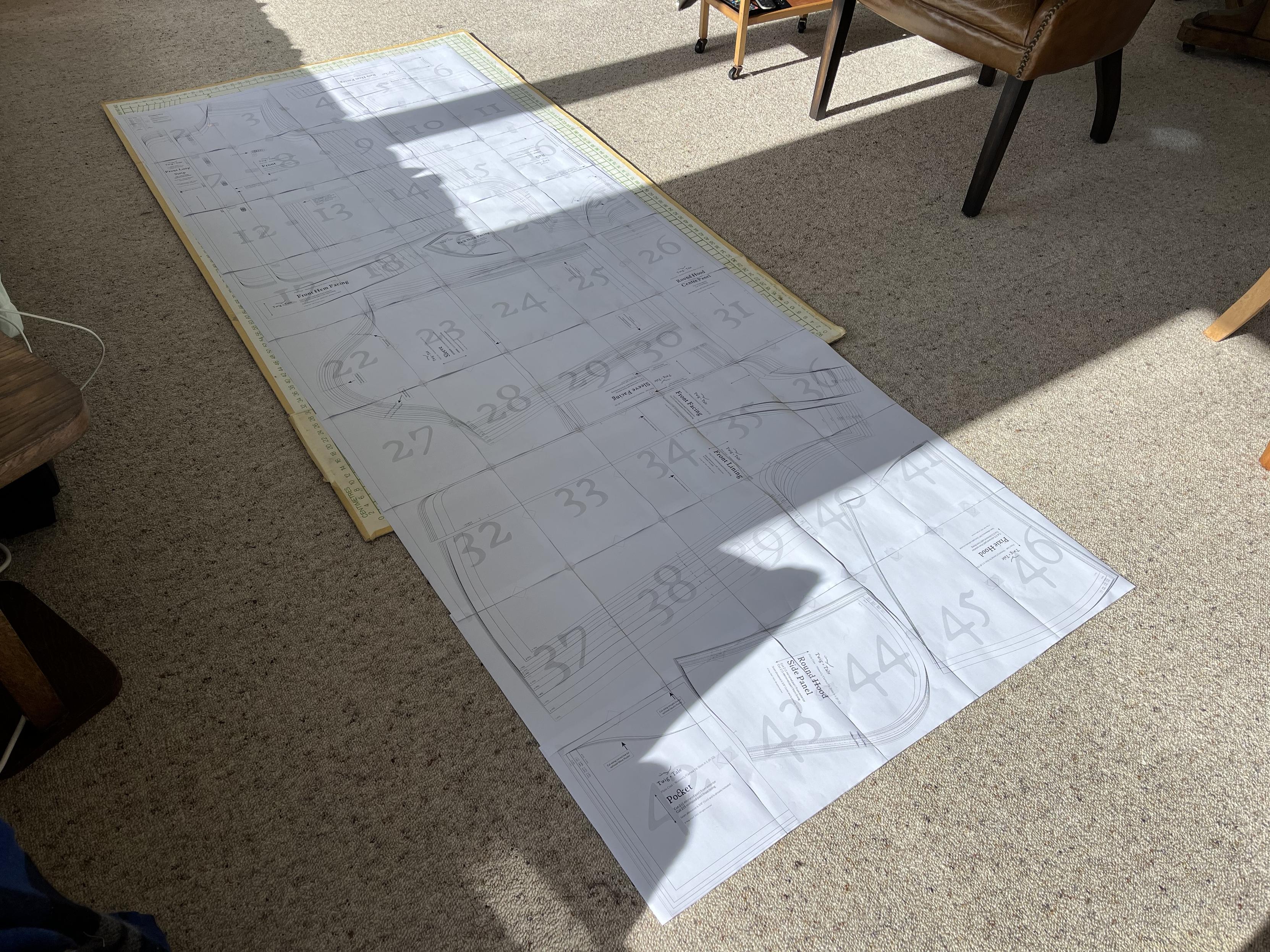Looking down on 45 pieces of A4 paper joined together on the floor, which is covered with fawn carpet. The edges of a cardboard cutting board are visible under the upper part of the paper. Printed on the paper are pattern pieces for the coat I’m going to make.