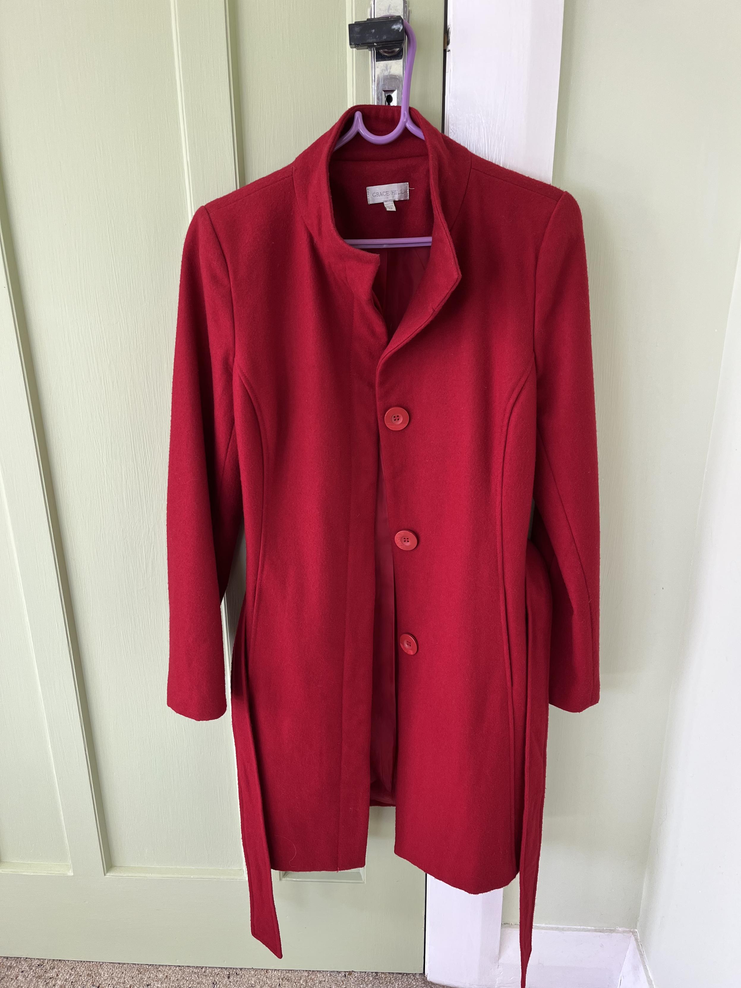 A bright red coat hanging on a coat hanger over a door knob. The coat is a slimline knee-length one with several panels, the seams highlighted with top stitching. It has a short stand up collar and large buttons down the front. The sash which ties around the waist is hanging at the sides.