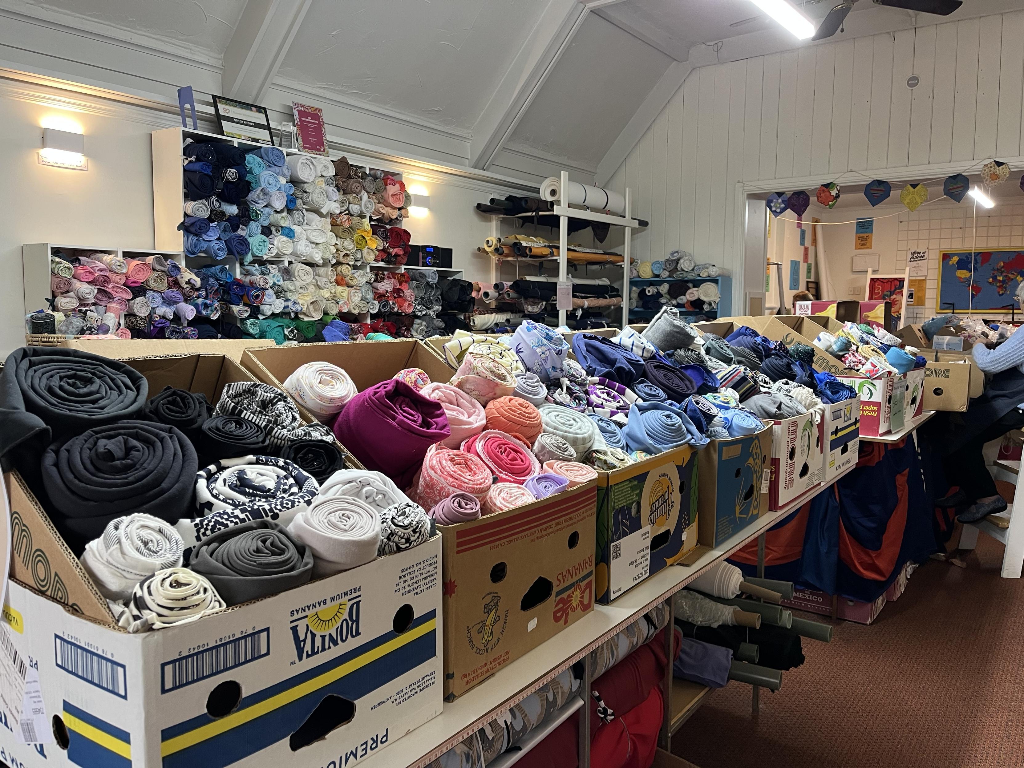 The interior of the Stitch Kitchen store, showing boxes of fabrics for sale. They are rolled up into bundles, sorted by fabric type and colour. In the distance are some longer rolls of fabric. Just visible at right is another room which is full of smaller offcuts for mending, crafts etc.
