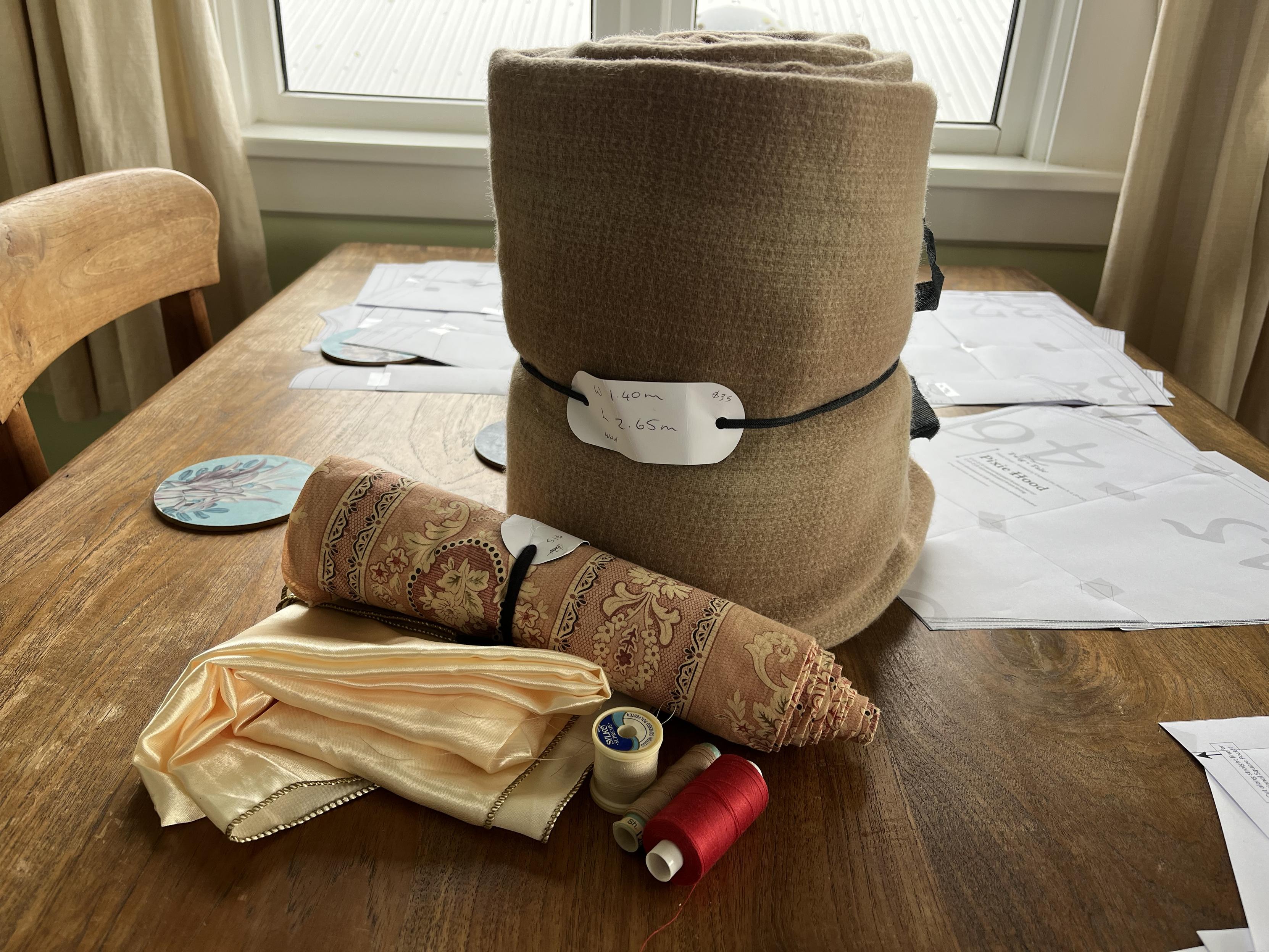 Sitting on a wooden table is a large bundle of beige woollen fabric. In front of it are a smaller bundle of fabric with a beige/pink design, some shiny cream lining fabric and 3 reels of thread. Also on the table are some paper pattern pieces.