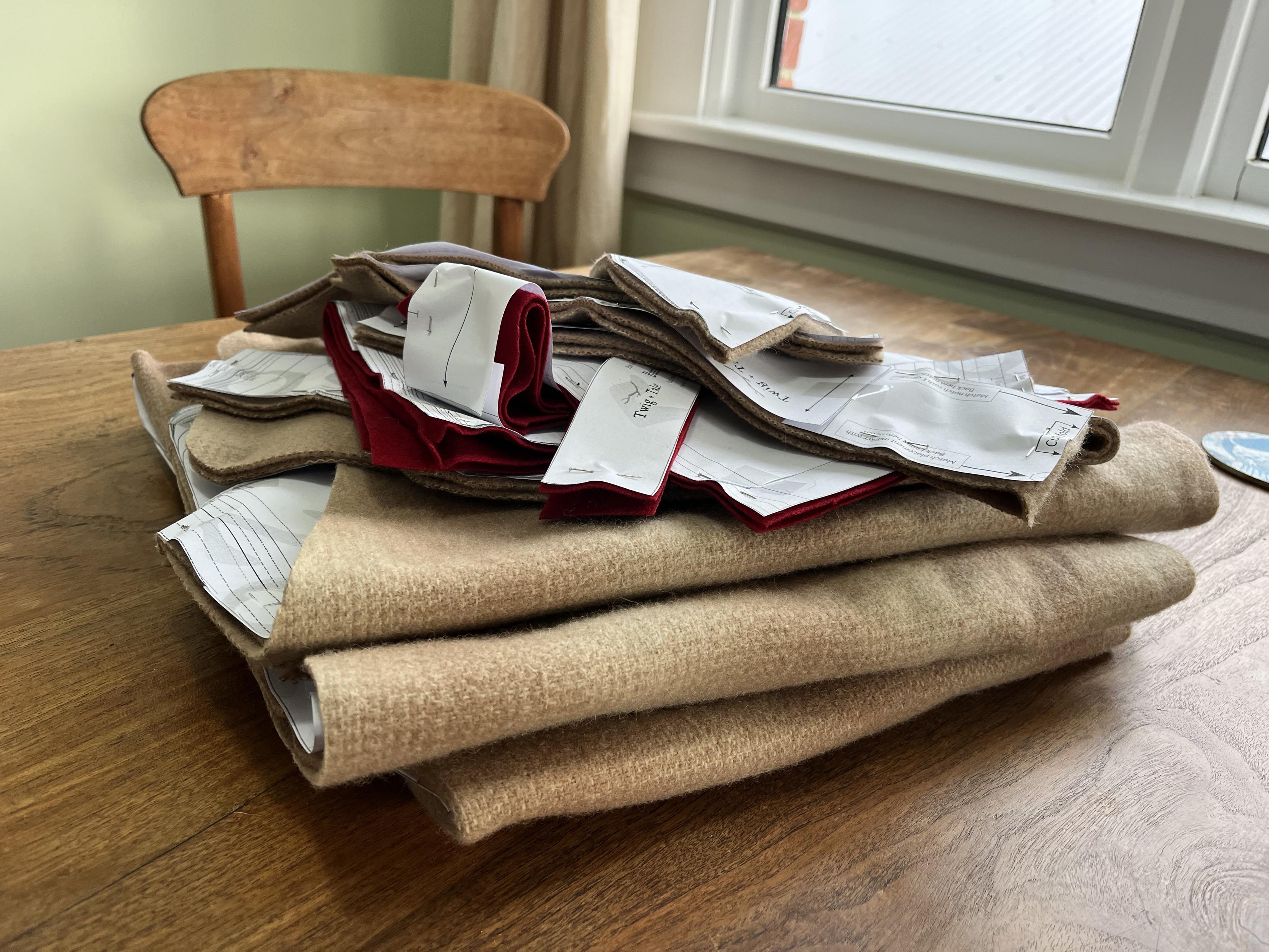 Sitting on a wooden table are a pile of folded pieces for a sewing project. The paper pattern pieces are still pinned to the fabric.Most of the pieces are in a beige/pink woollen fabric with a faint check pattern. A few are in a plain red fabric.