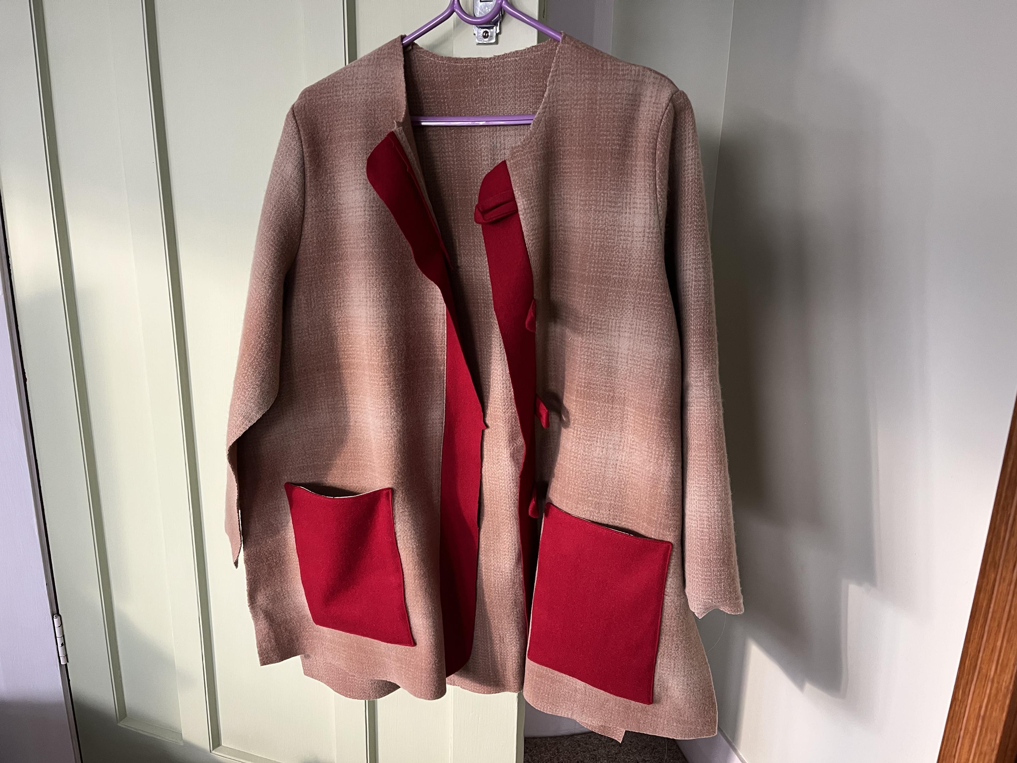 A partially-made coat, on a hanger on a door handle. The main fabric of the coat is a pink/beige wool with faint check pattern. The pockets and front button strips and button loops are red. The back, fronts and sleeves are sewn together but the sides aren’t stitched together yet.