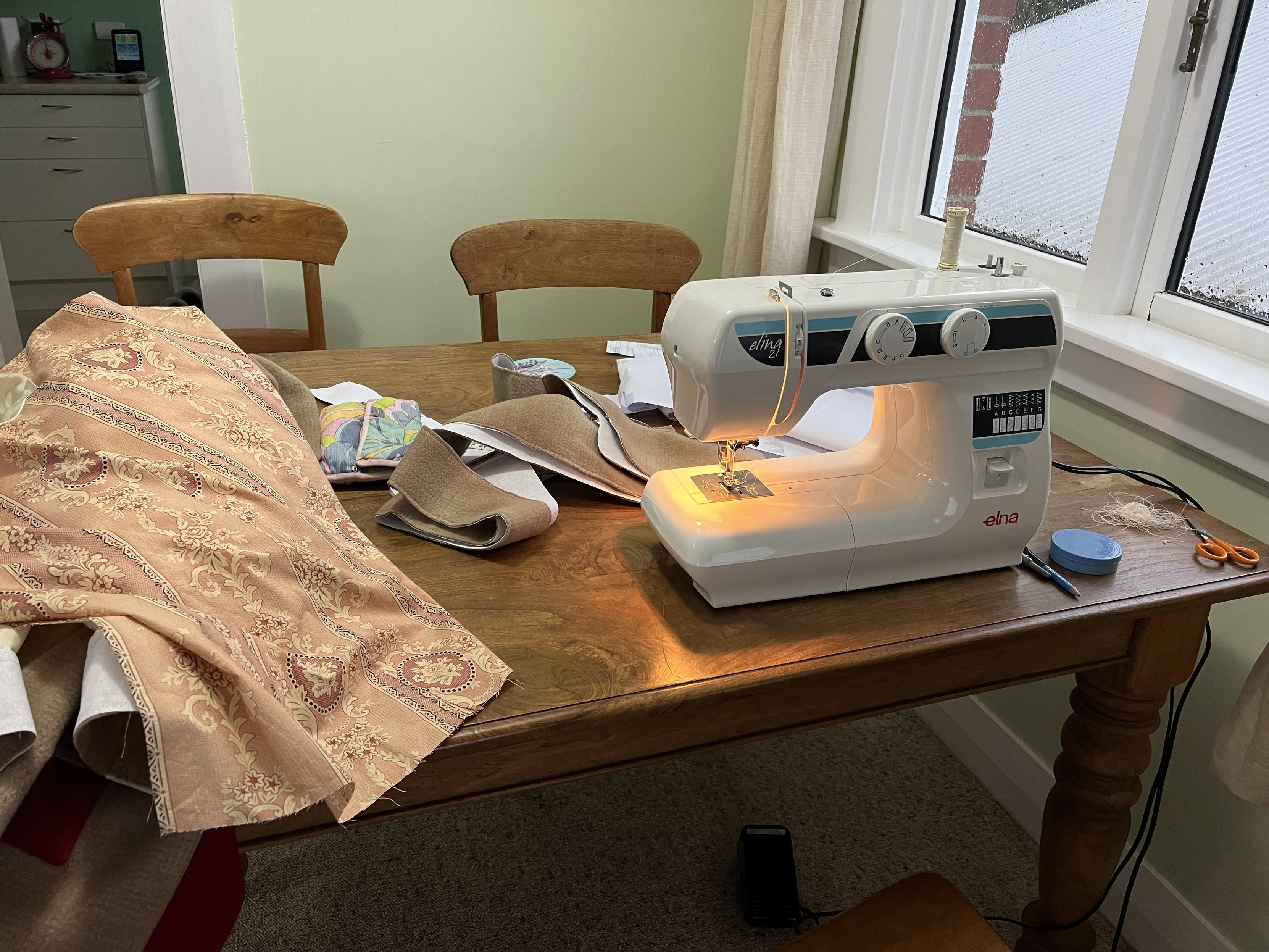 Sitting on a wooden table is an Elna sewing machine, with a yellow glow coming from its built-in light. Also on the table are various sewing necessities, including scissors, unpicker, tin of pins, and a small quilted sewing bag. There are bits of fabric, including a large one in beige/pink floral design - that’s the lining for the coat I’m making.
