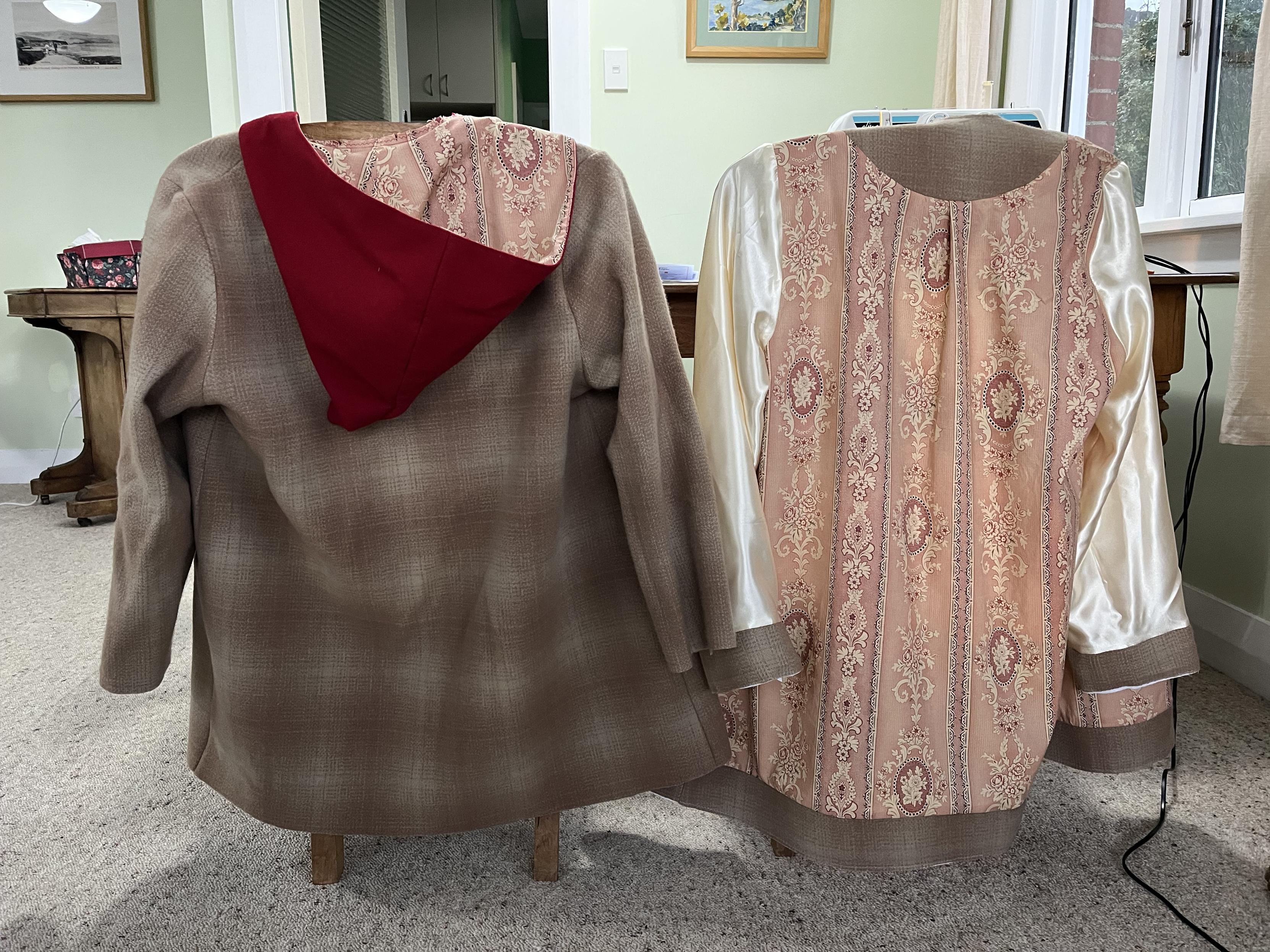 Draped over the backs of chairs are the 2 parts of the coat I’m making. On the left is the exterior, made of a pinky beige woollen fabric. It has a red hood, lined with a pink floral/geometric cotton. On the right is the lining of the coat. Most is in the same fabric as the hood lining, but the sleeves are plain cream. Facings are attached to the lining. Not visible are the red pockets and button plackets on the front of the coat.