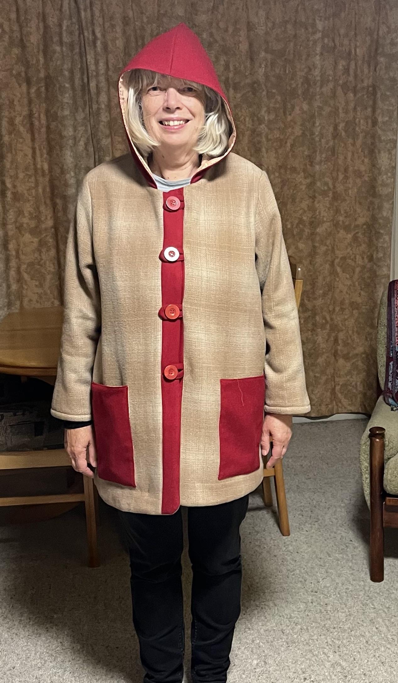 Me, a middle-aged white woman with a grey bob and fringe, modeling my new coat. It’s thigh-length and has a pixie hood. The main fabric is a beige faintly checked wool fabric, with contrasting red fabric for the pockets, button strip and hood.