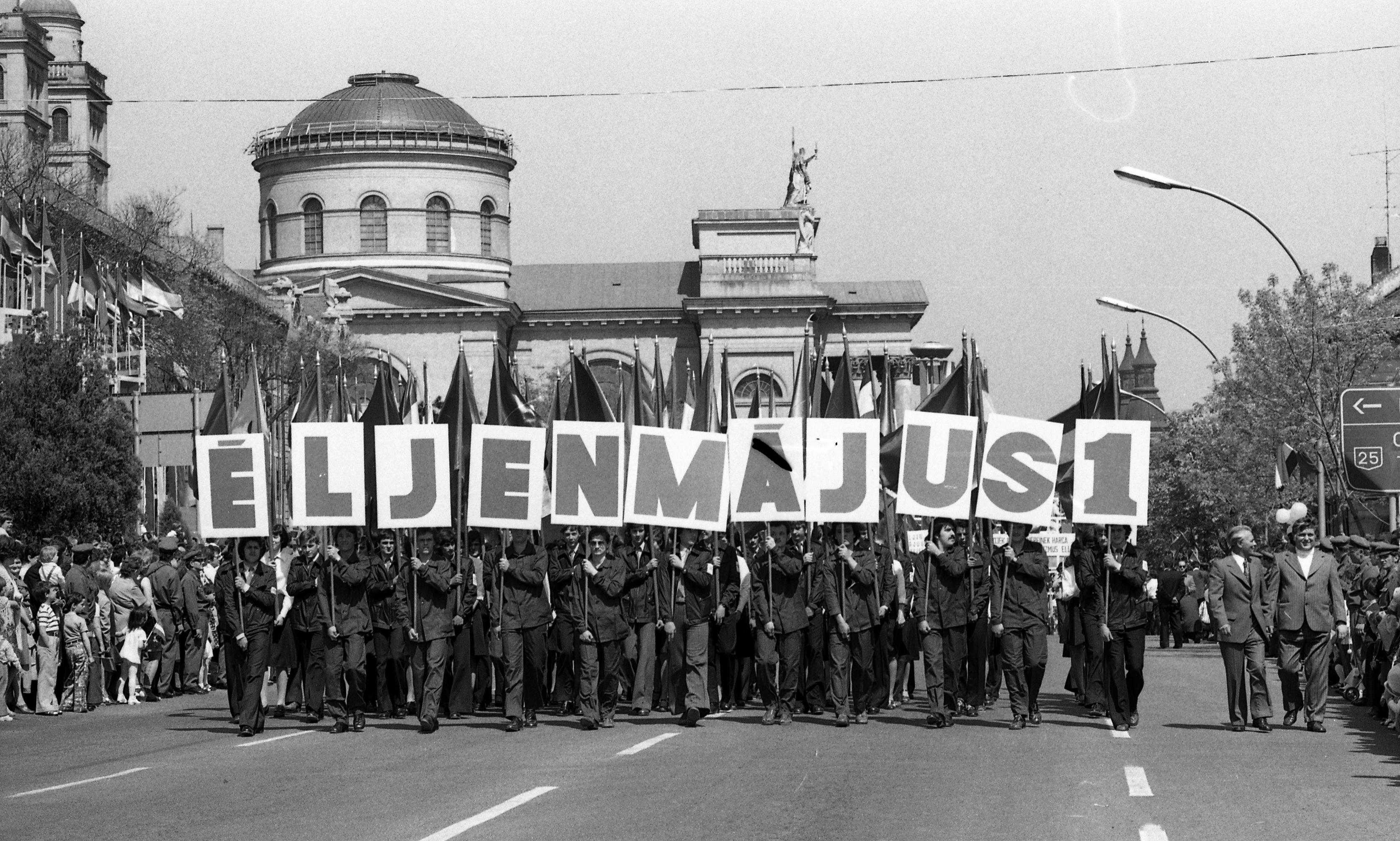 A black and white photo of a group of people marching on a road with those people in front holding up a sign with single letters that spell out Éljen Május 1 or 'Long Live the 1st of May. People behind the front row are carrying large flags.