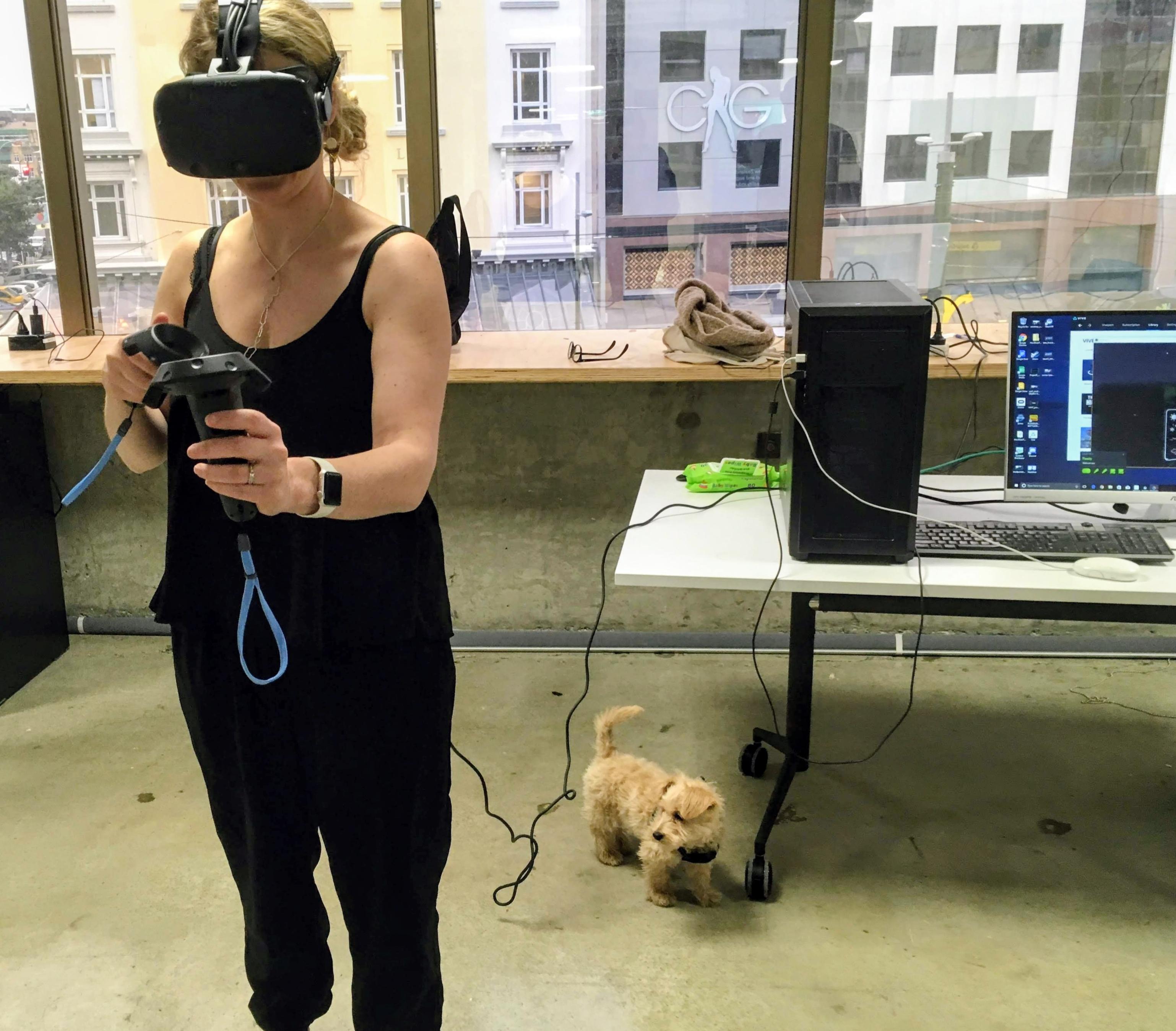 An adult with blonde hair wearing a black tank top and black trousers uses an HTC Vive VR headset and controllers in an office setting. They are standing in the center of the room, while a small, scruffy, tan-colored puppy stands near their feet, looking toward the camera.

In the background, a large window reveals a city street with multi-story buildings. To the right, a white desk holds a black PC tower, a monitor displaying a VR interface, a keyboard, and a mouse. A few personal items, including a pair of glasses and a small cloth, sit on the windowsill.