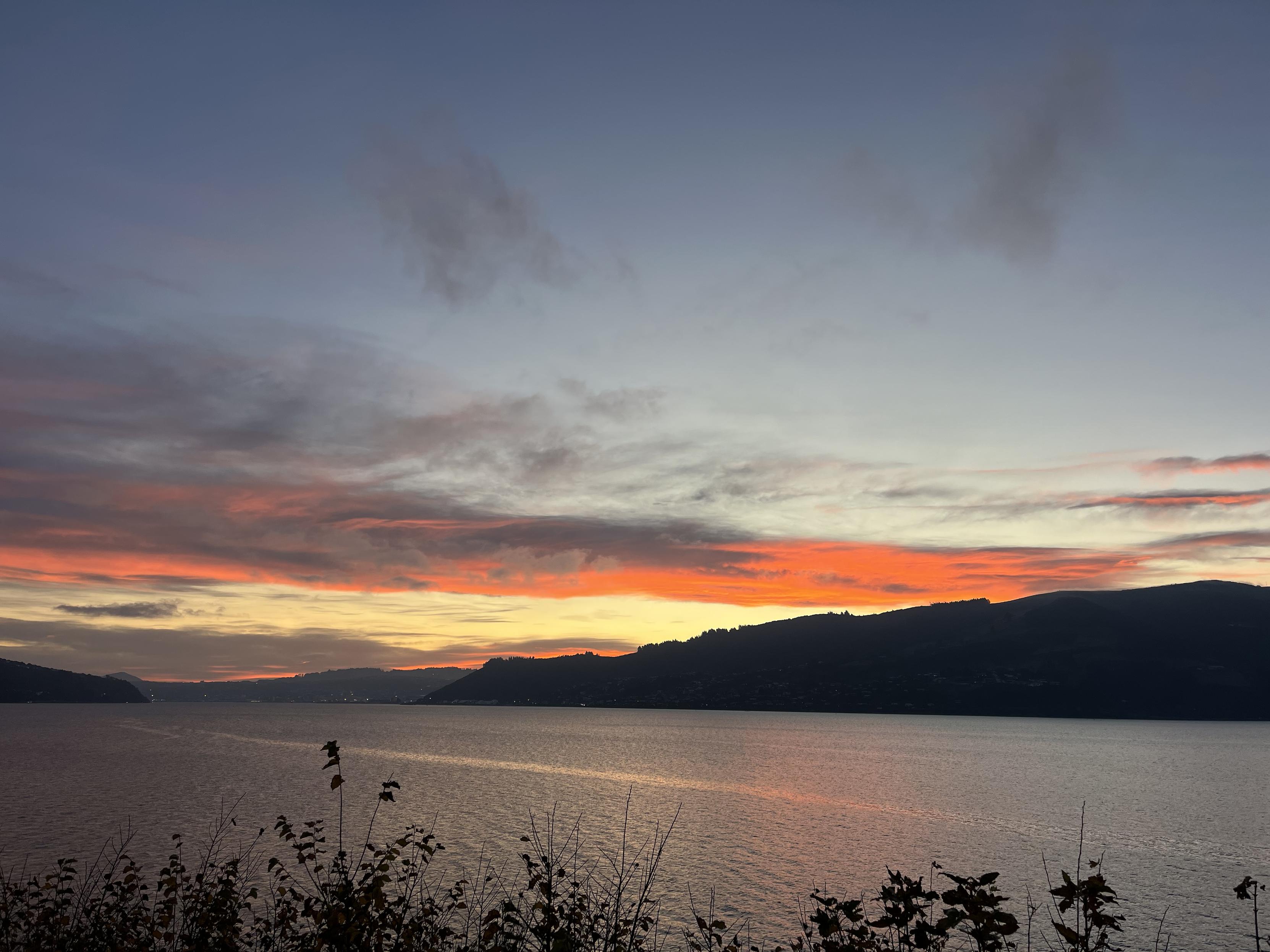 Looking across Otago Harbour from the peninsula at dusk. It’s partly cloudy and the clouds are a neon orange shade from the sunset.