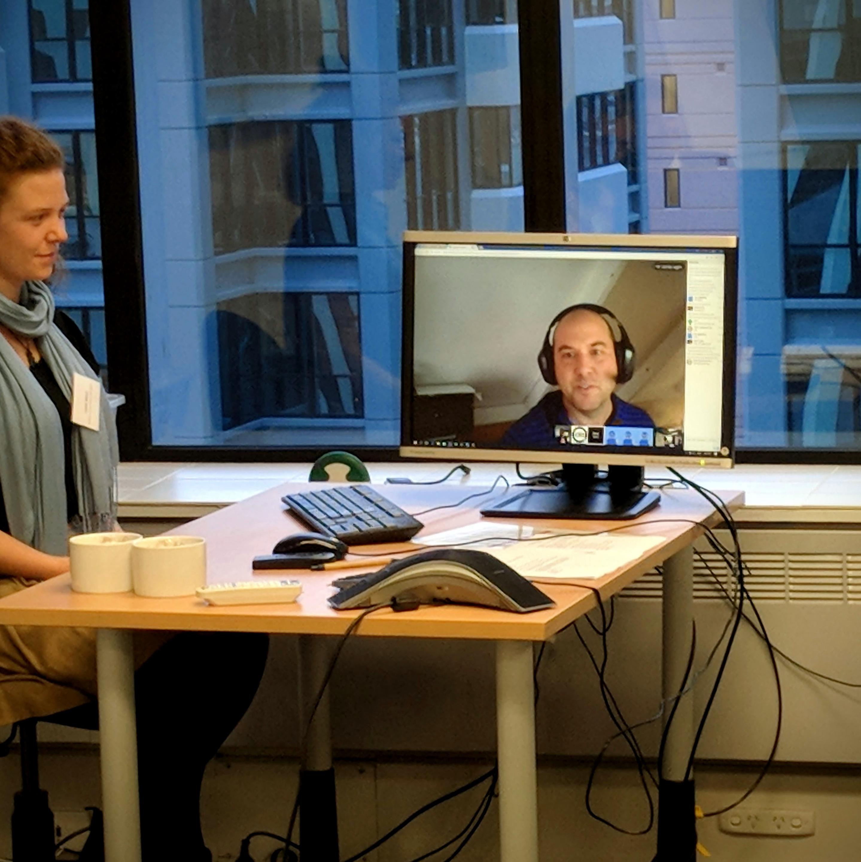 A professional Q&A session with the NZ Privacy Commissioner, captured via video call in 2018. The computer monitor prominently features a man with a headset participating from a remote location. In the foreground, a woman sits at an office desk equipped with a conference speakerphone and two white mugs, looking toward the screen. Large windows behind the desk reveal the blue exterior of a neighboring city building, framing the digital meeting.
