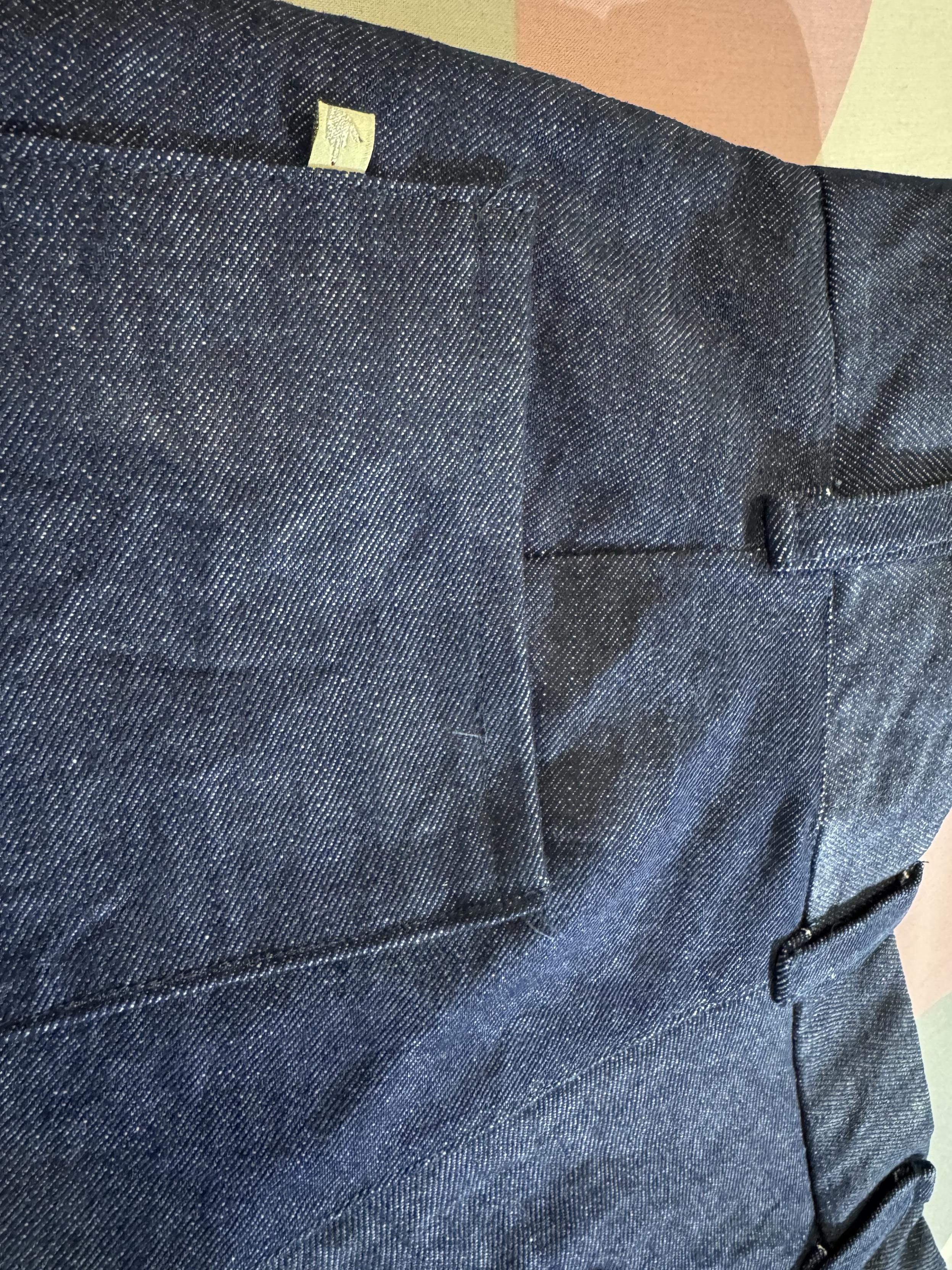 Back view of a pair of blue denim shorts with a patch pocket, belt loops and a small gold coloured label