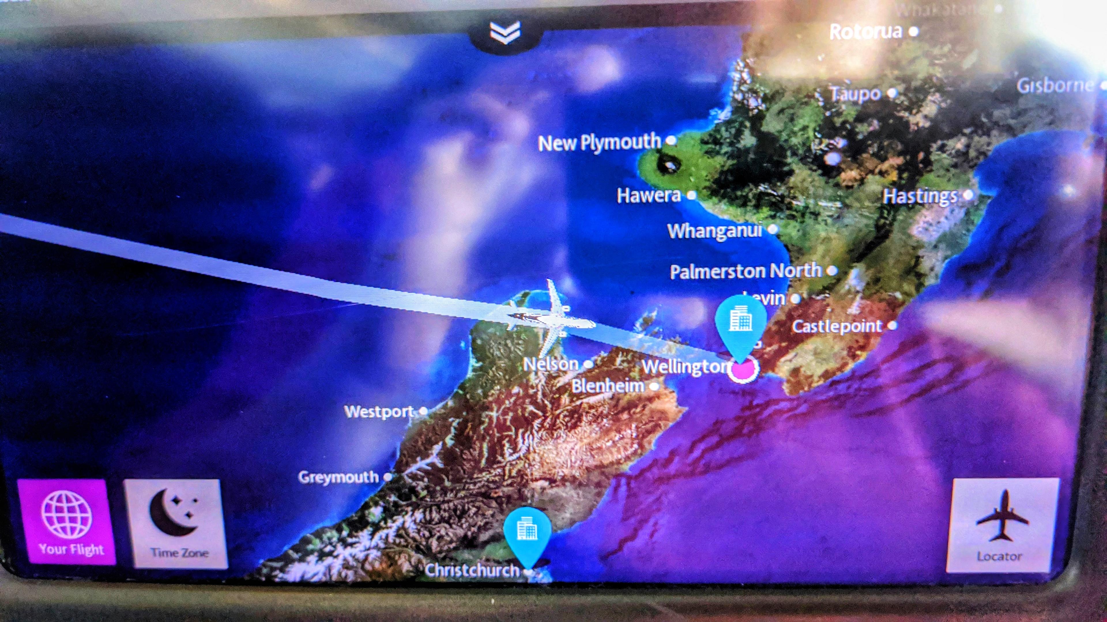 A photograph of an in-flight entertainment screen showing a digital flight map of New Zealand. A white airplane icon is positioned over the Cook Strait, heading toward Wellington. The map highlights various cities including Nelson, Christchurch, Palmerston North, and Hastings against a green and brown topographical landscape surrounded by deep blue ocean. Bright pink and blue location pins mark Wellington and Christchurch. In the corners of the screen, touch-sensitive icons for "Your Flight," "Time Zone," and "Locator" are visible.