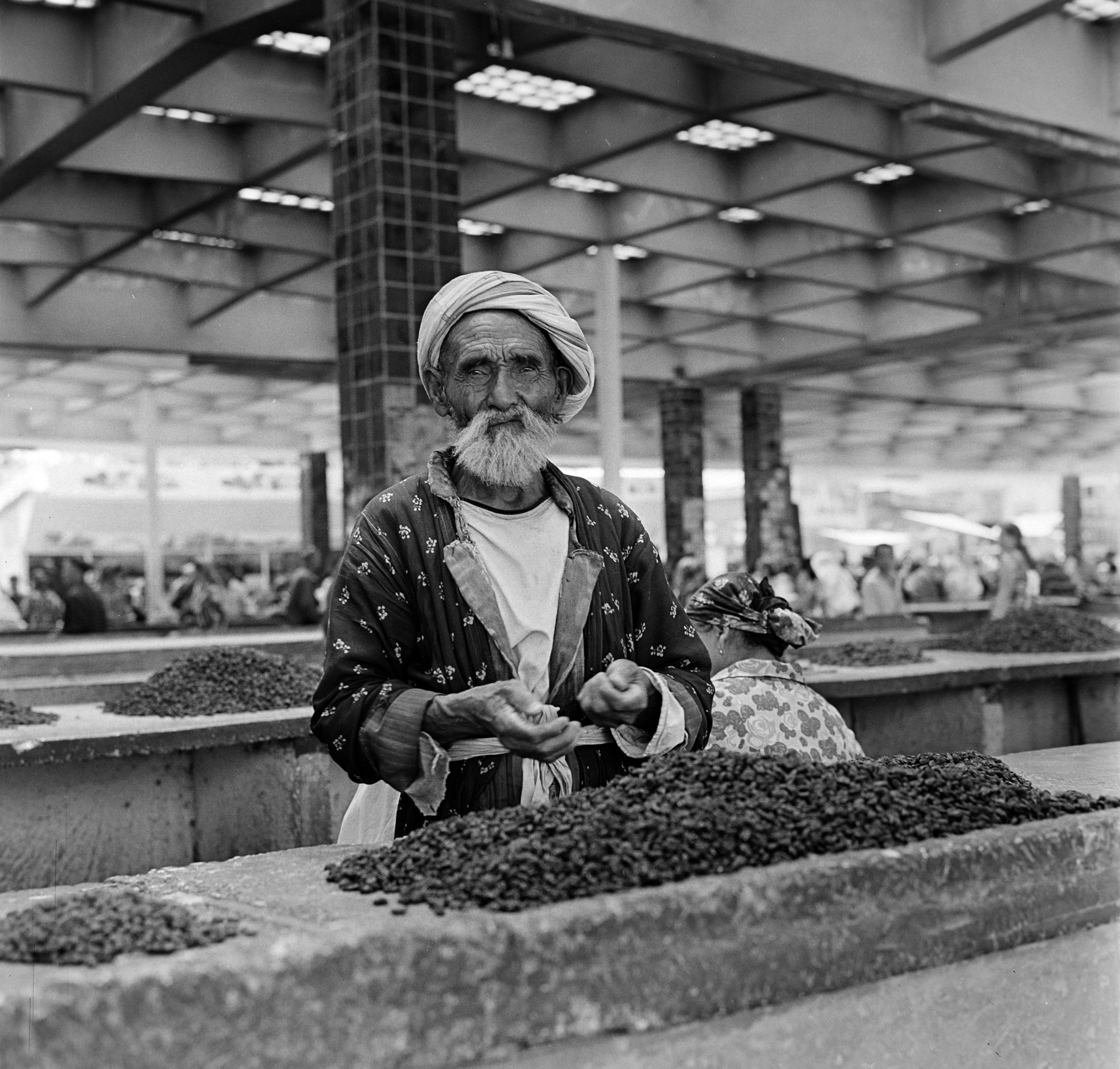A black and white photo of an old man standing at a counter with a large amount of what appears to be beans in front of him.  He is in large market hall and there are other counters or benches, also with piles of beans on them. The old man has a largish, white beard, is wearing an open pattern jacket, tied with cloth, and a turban.
