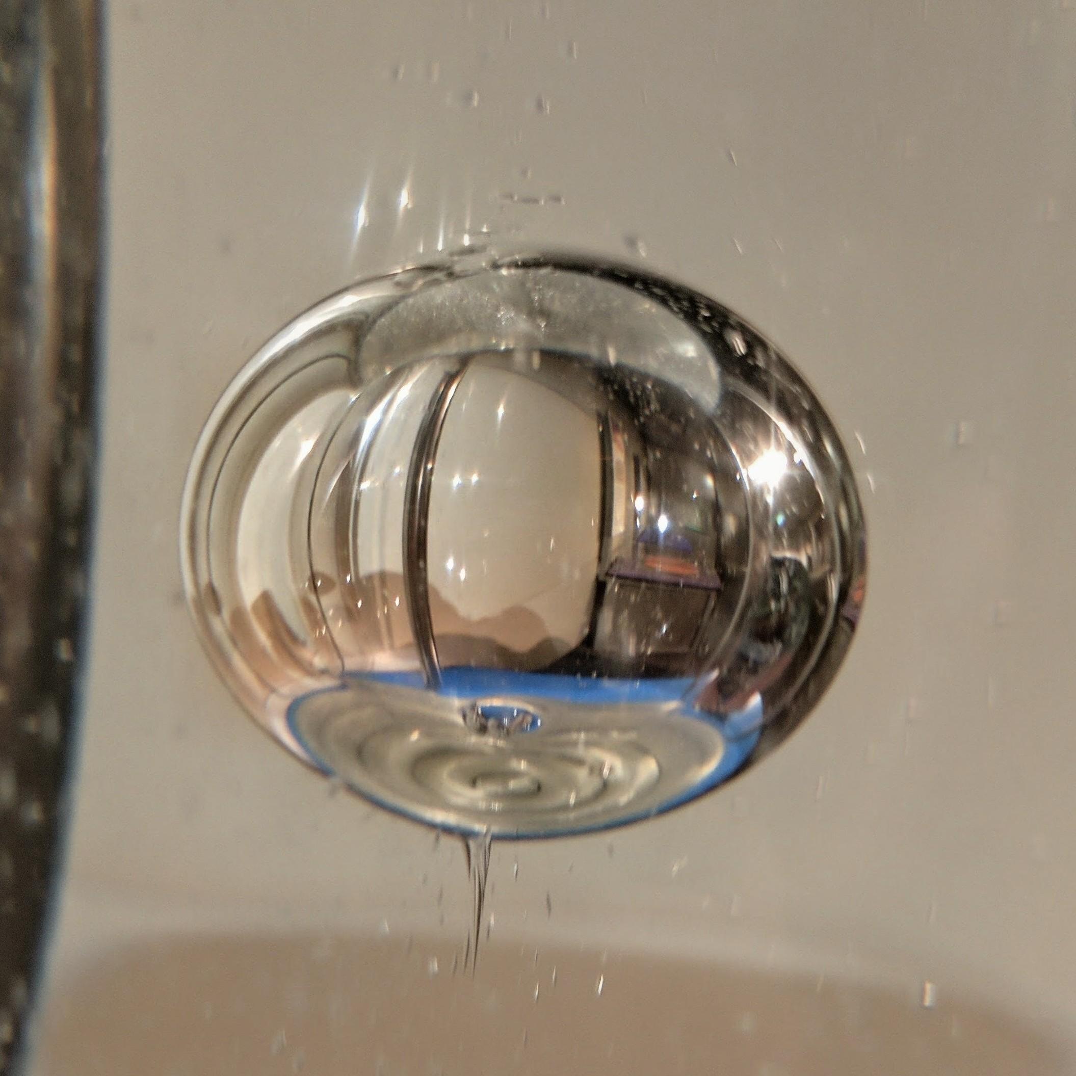 A close-up photograph of a large, spherical air bubble floating underwater. The bubble acts like a fisheye lens, showing an inverted reflection of a room featuring light walls and a bright blue surface. A thin, watery trail extends downward from the bottom of the bubble, and tiny, out-of-focus bubbles drift in the surrounding water.
