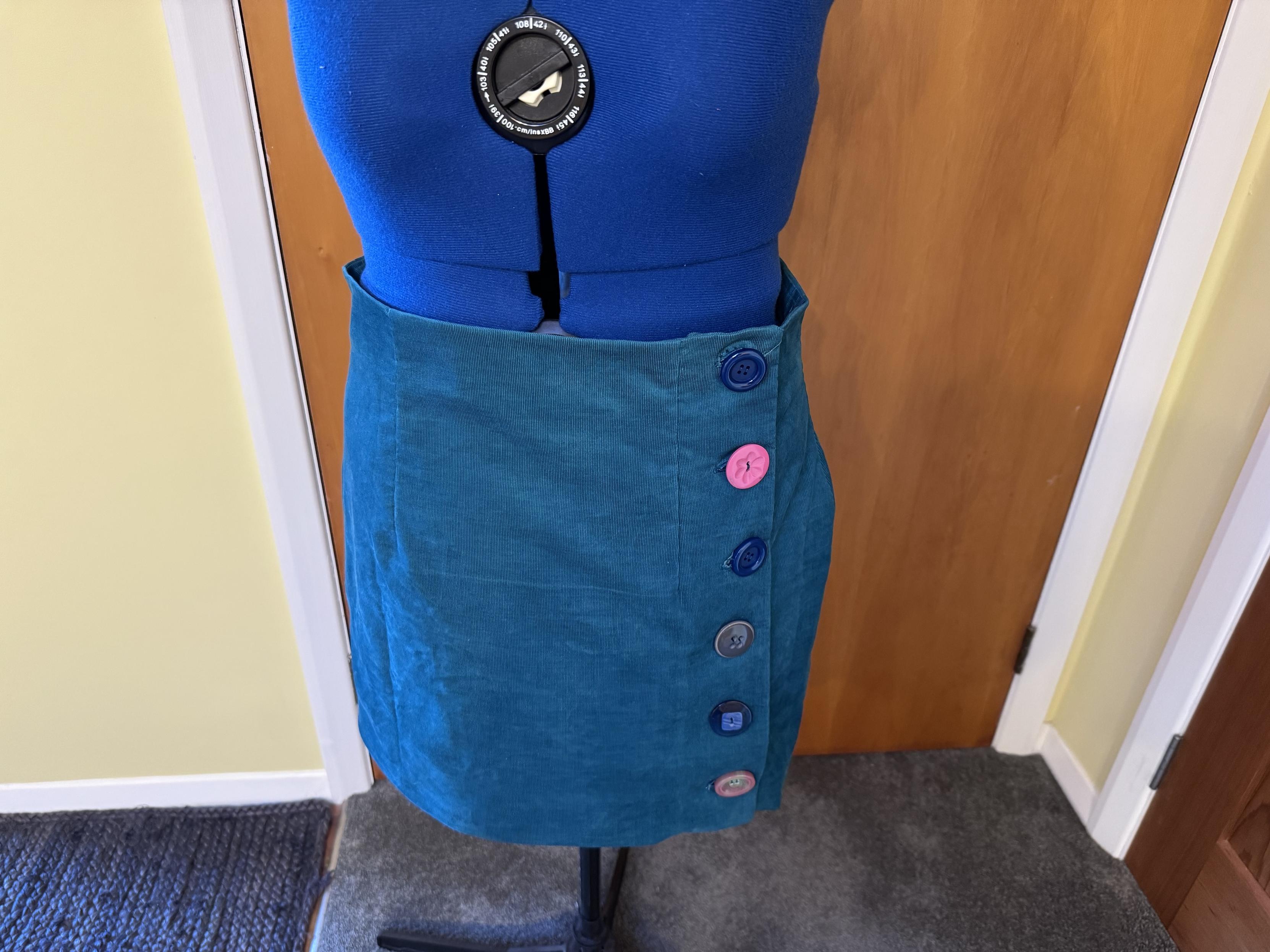 A teal skirt displayed on a dress form, with a row of colorful buttons on one side. The background features a wooden door and a carpeted floor.