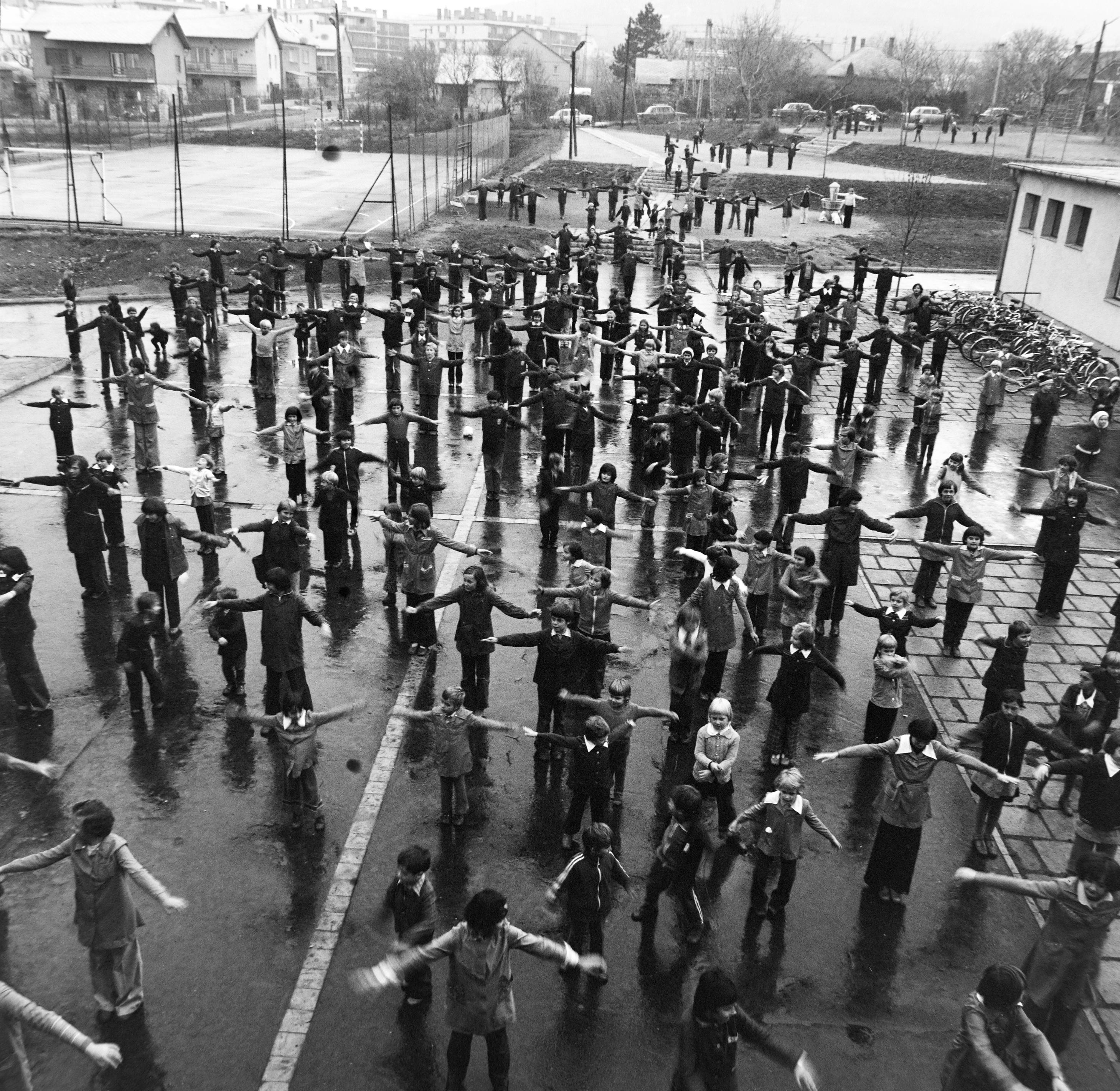 A black and white photo of a large number of children doing what looks like physical education exercises on a wet school playground.  Most of the children have the arms stretched out to their sides but some don't.  They all look a bit all over the place.  In the distance there are flats and houses.