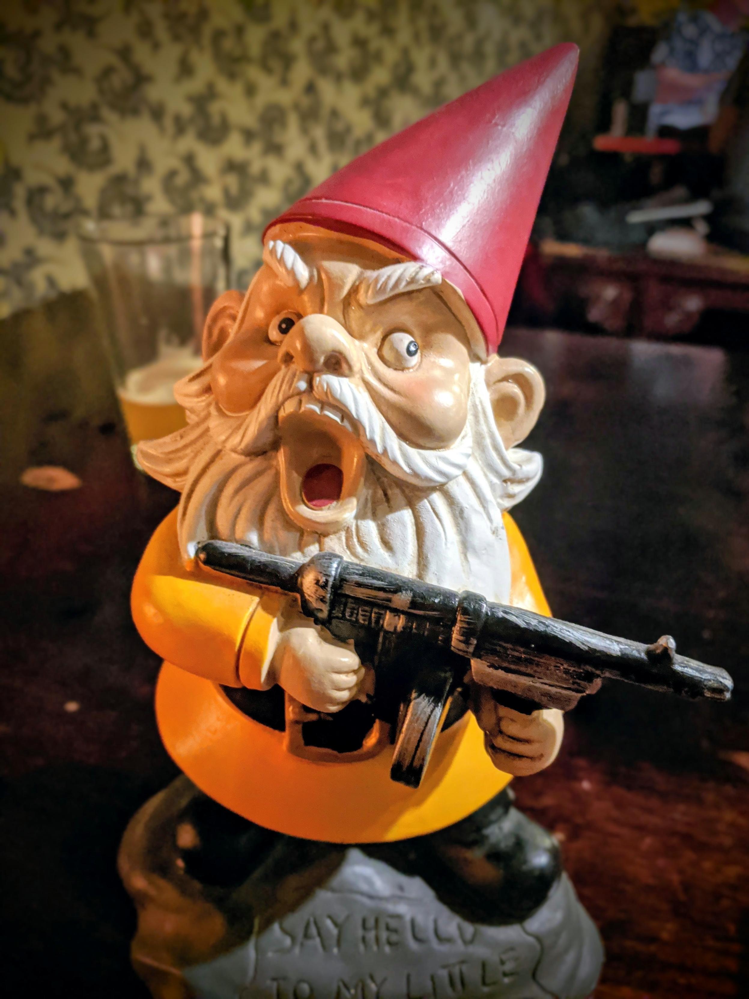 A close-up shot of a novelty garden gnome figurine with an angry, yelling expression and an open mouth. The gnome wears a classic pointed red hat, a yellow tunic with a black belt, and has a long white beard. It is aggressively holding a black submachine gun. The grey rock base of the figurine features the inscribed text, "SAY HELLO TO MY LITTLE FRIEND". The background is a softly blurred indoor setting with a pint glass of beer on a dark wooden table.