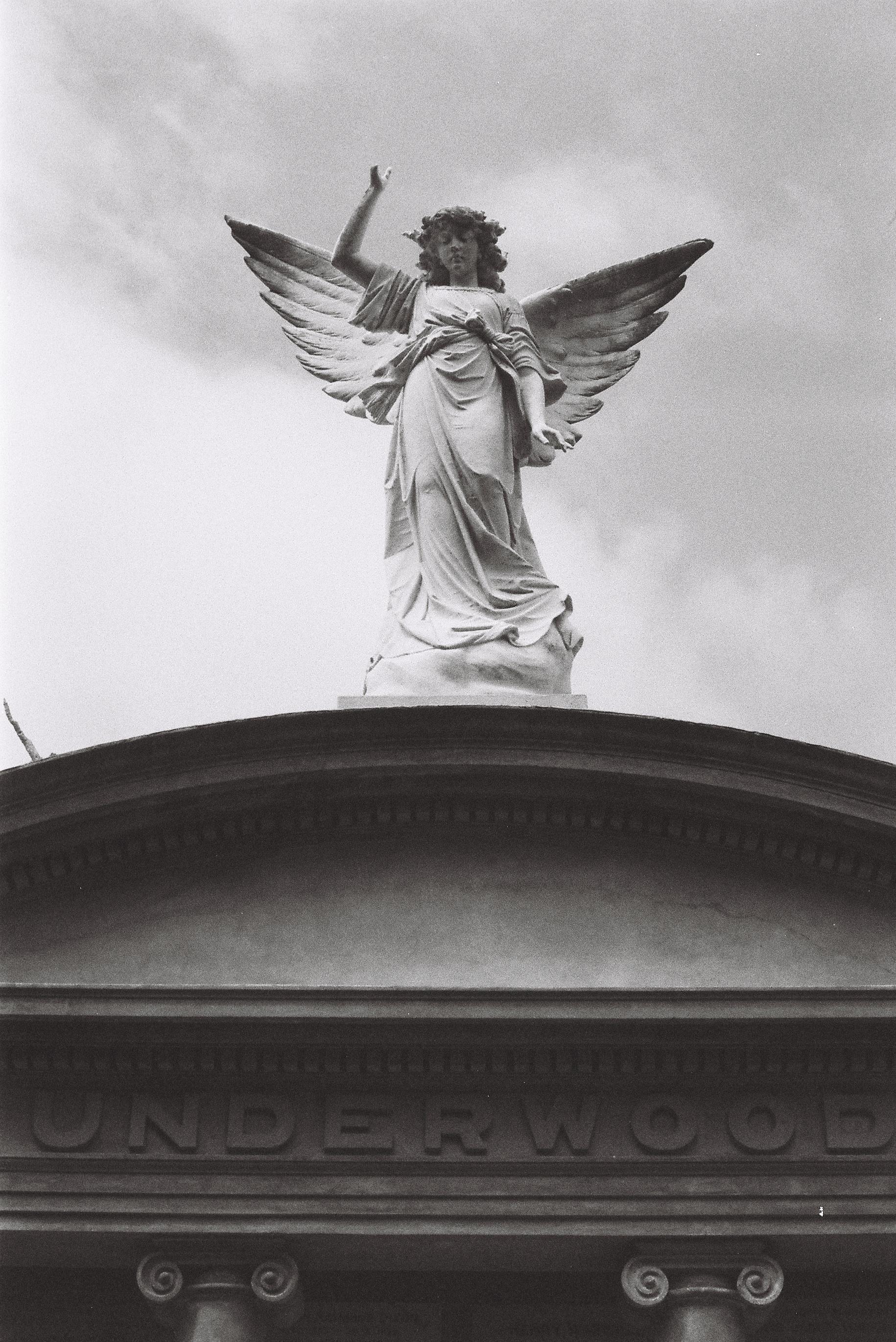 A vertical portrait black & white photo of an angel with spread wings and one hand held aloft standing on the curved stone roof of a family tomb. The family name 'Underwood' is carved into the stone below the roof eaves.
Photo was shot on a Pentax MX camera & Fomapan 400 film