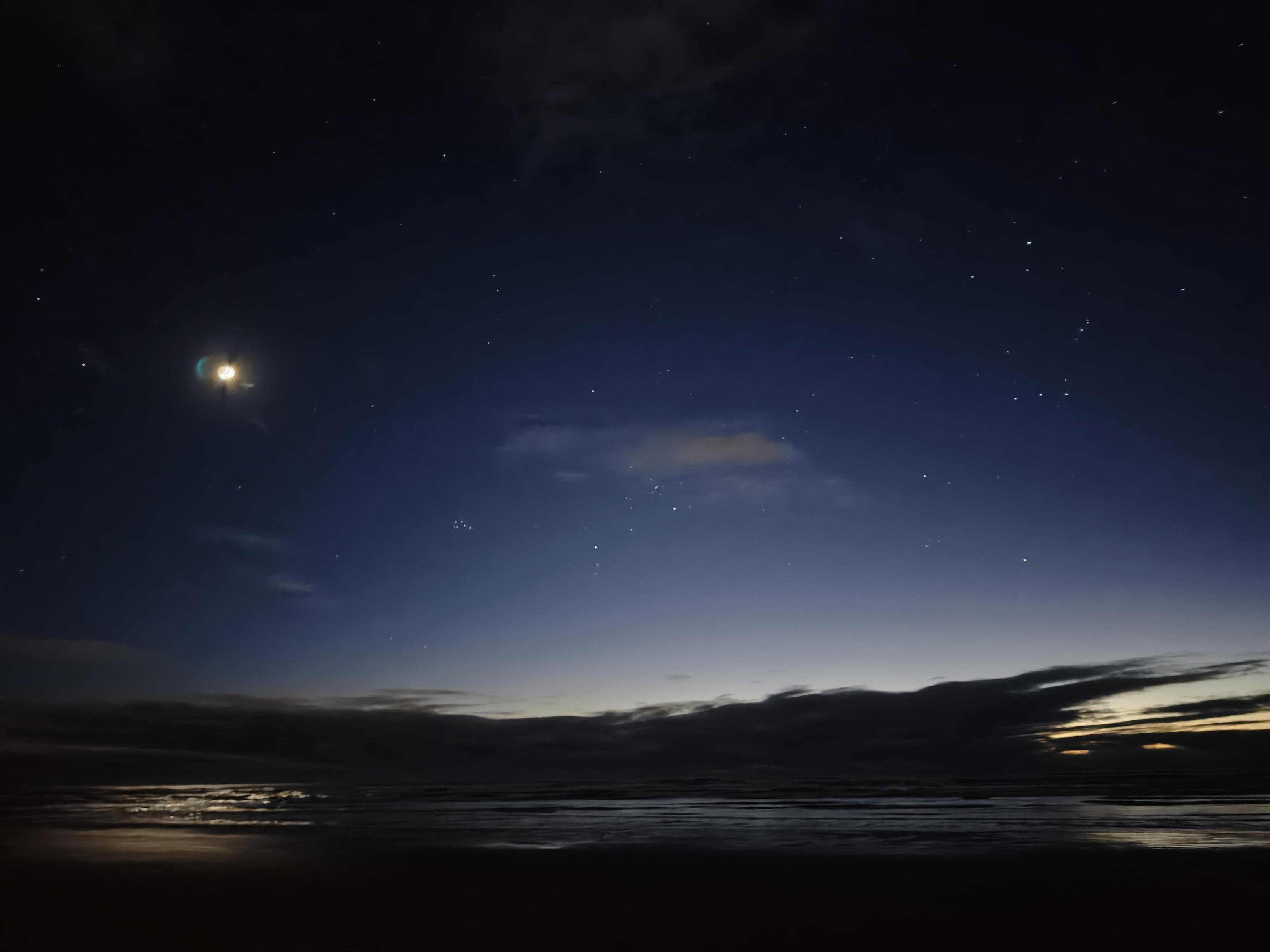 A photo of the very early morning sky above the beach, mostly dark and star-studded with the morning light starting to bleed in over the horizon. The moon is on the left side of the frame, and the constellation Orion is on the right. Between them, centre left, is Mātariki, a dense cluster of stars.