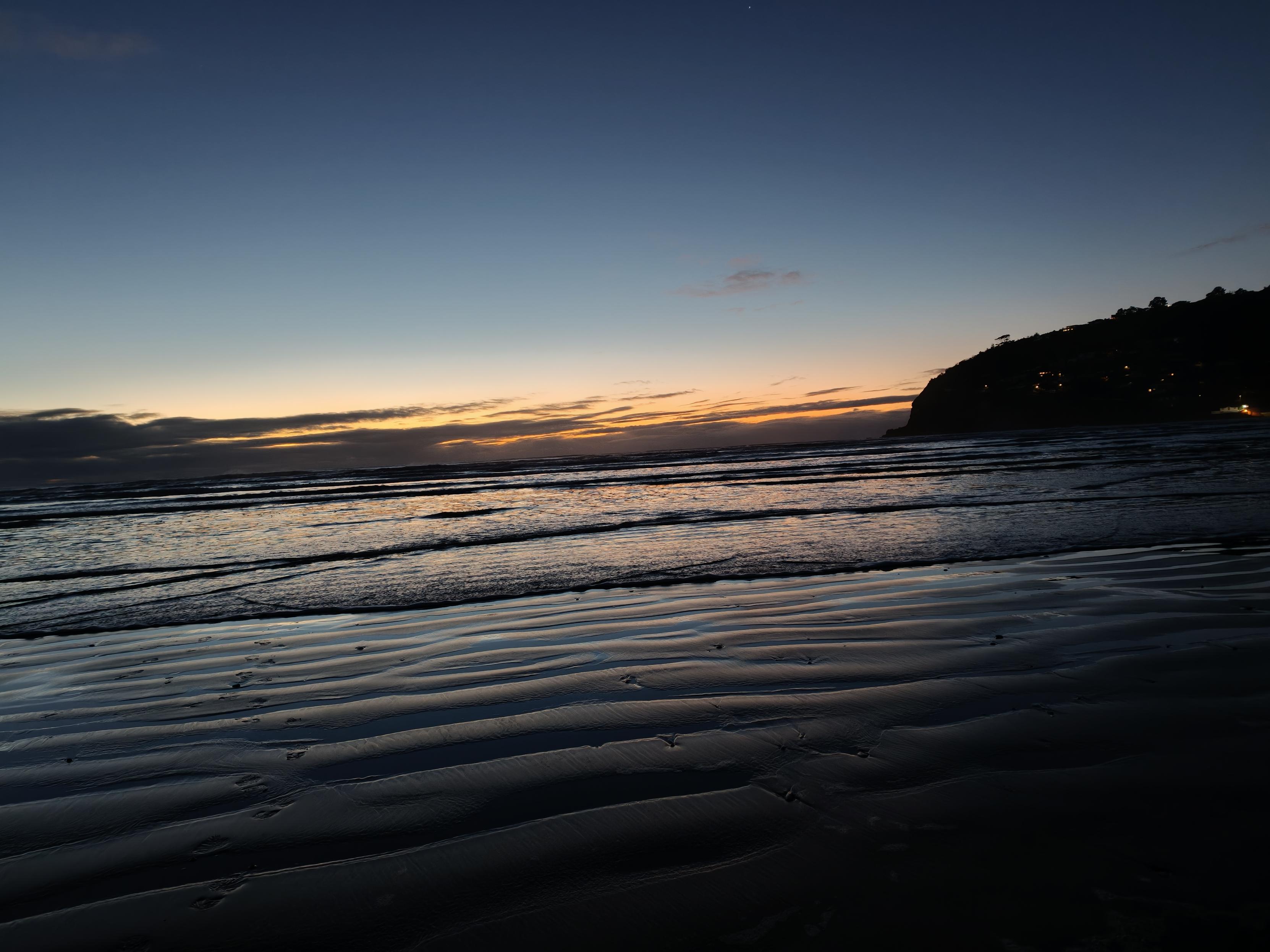 A photo of the same beach but the sun is starting to rise, the sky lighter blue and orange. The wet sand and an oncoming wave reflect the gradient.