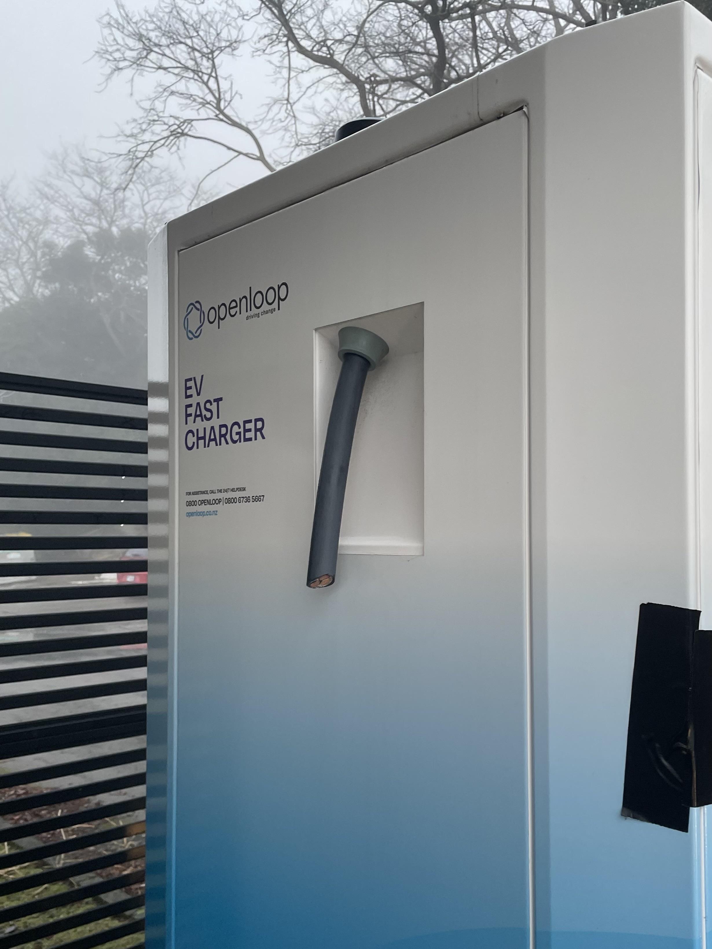 The side of a white and blue OpenLoop EV fast charger. A large thick black cable comes out the side, and is abruptly cut off. There is no cable or charger attached to this machine.