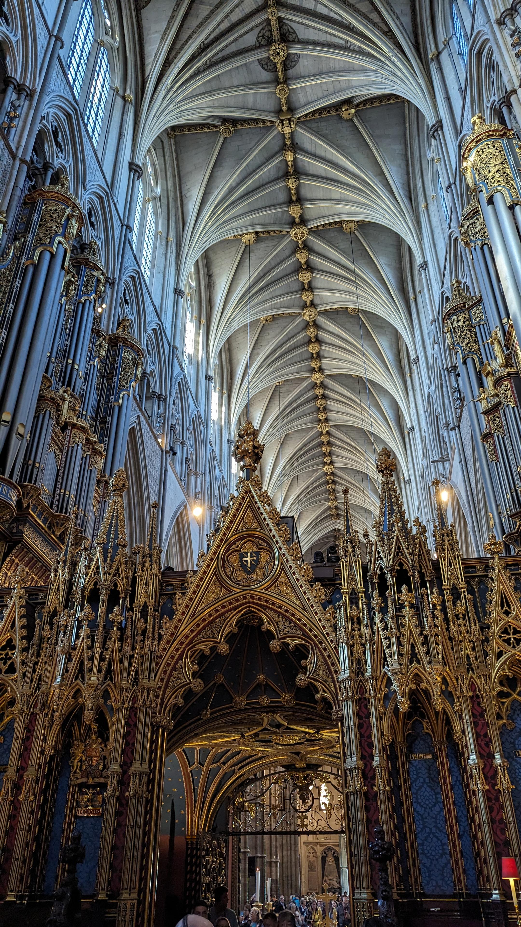 An interior photograph of Westminster Abbey, taken from the nave looking east toward the high altar. The view captures the immense scale and intricate details of the Gothic architecture, specifically the elaborate gold-covered choir screen in the foreground and the soaring, ribbed stone vaulting of the ceiling above. Tall clerestory windows line the upper walls, and a few visitors are visible standing at the far end of the nave, emphasising the building's height.