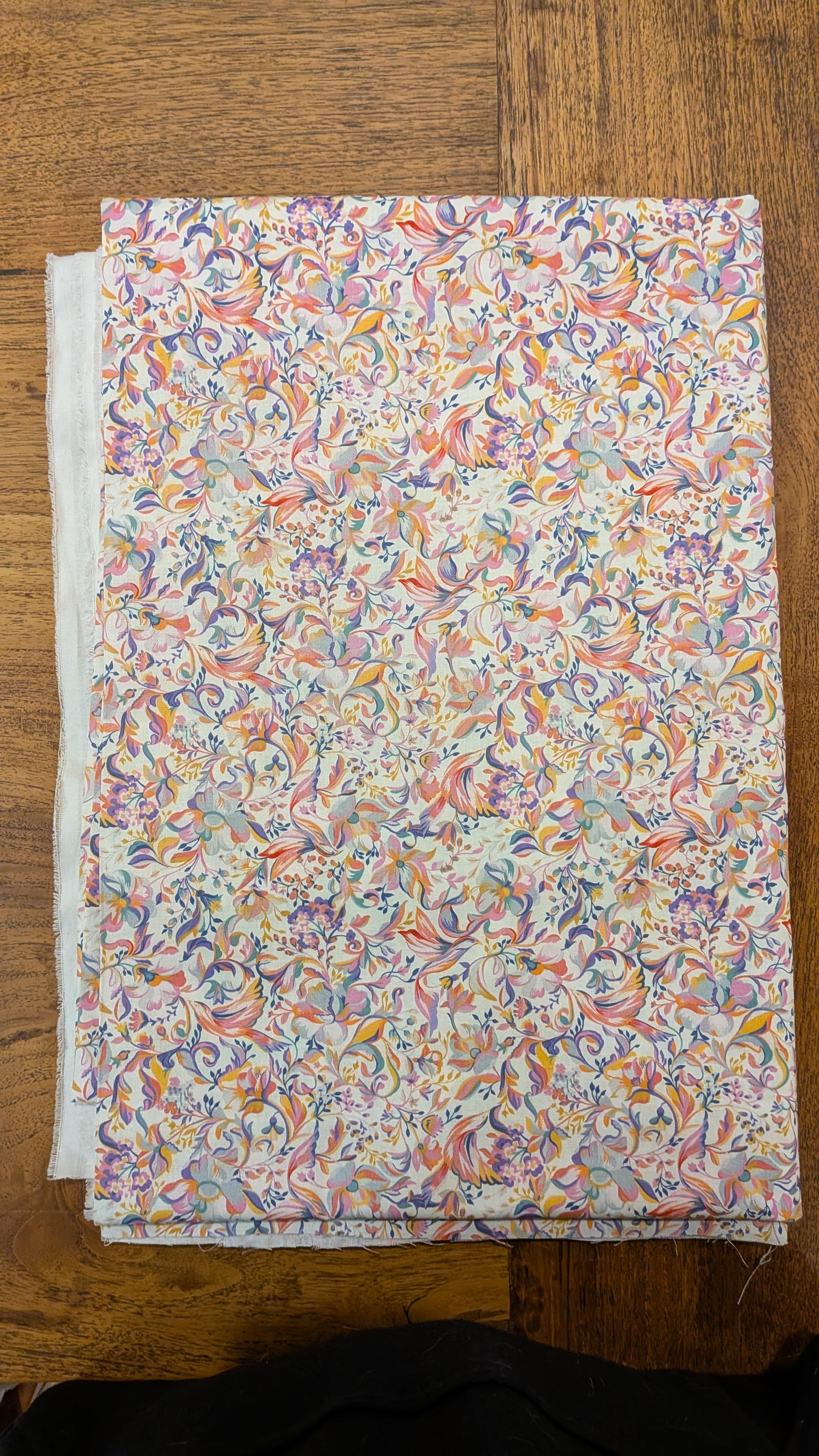 a photo of fabric on a wooden table. the fabric has a white background with a pink, orange, blue and purple sort of floral curlicue/art nouveau/florentine design