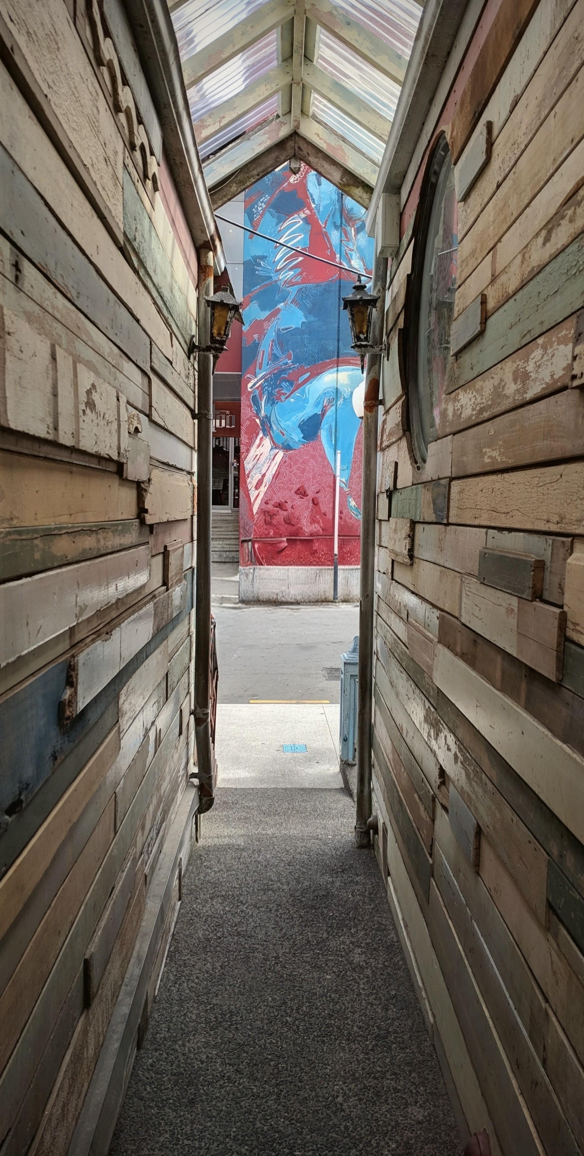 ​A view from inside the entrance of the Havana Bar in Wellington, looking out onto Wigan Street. The perspective is framed by a narrow, covered walkway lined with weathered, multi-colored horizontal wooden planks and topped with a glass gabled roof. At the end of the walkway, a large, vibrant, abstract mural in red and blue is visible on the building across the street.