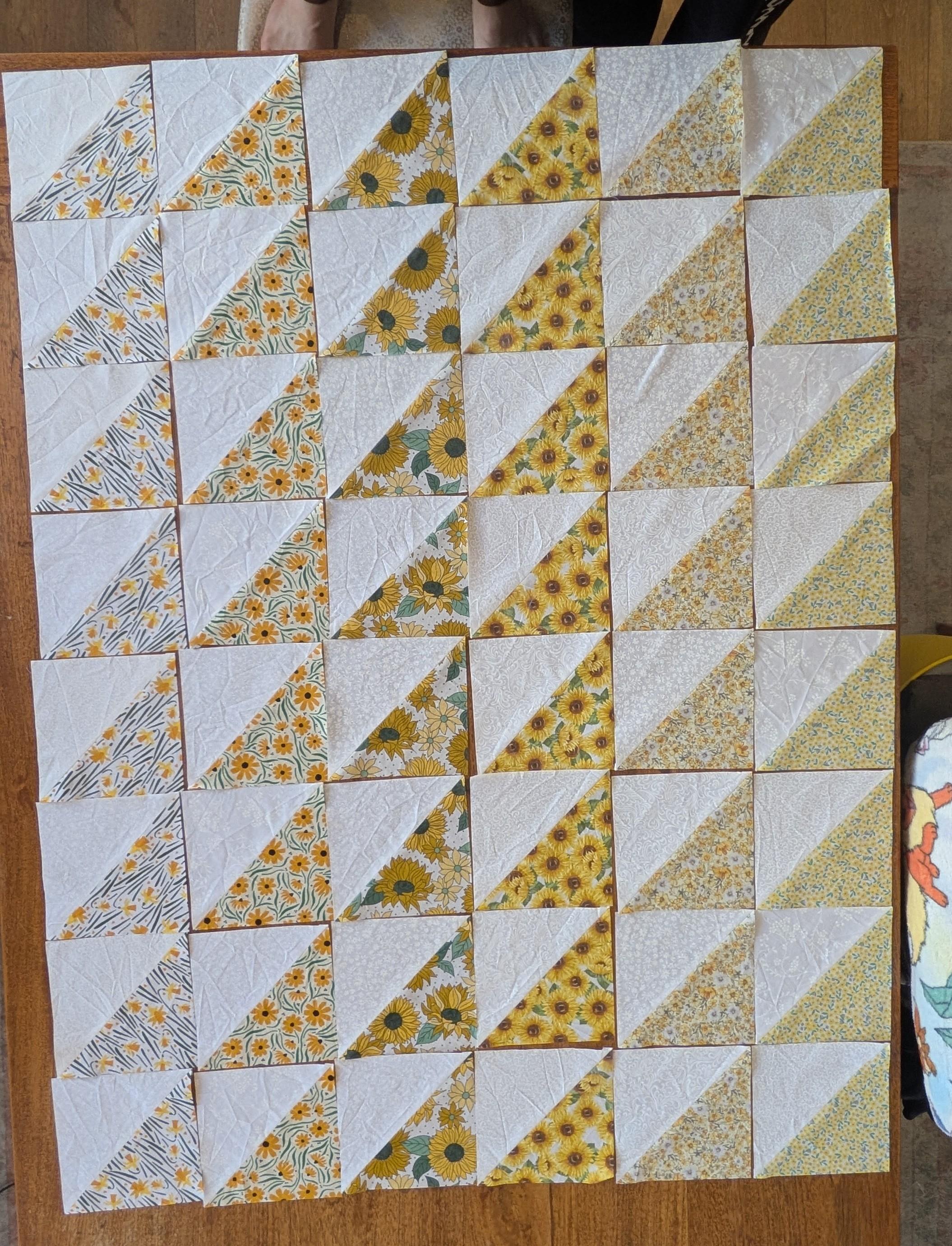 top-down shot of a quilt top, a 6 x 8 grid of quilt blocks laid out on a wooden table. each block is half white fabric, half yellow floral across a diagonal line. they're roughly arranged in order colour order, starting darker yellow and trending lighter