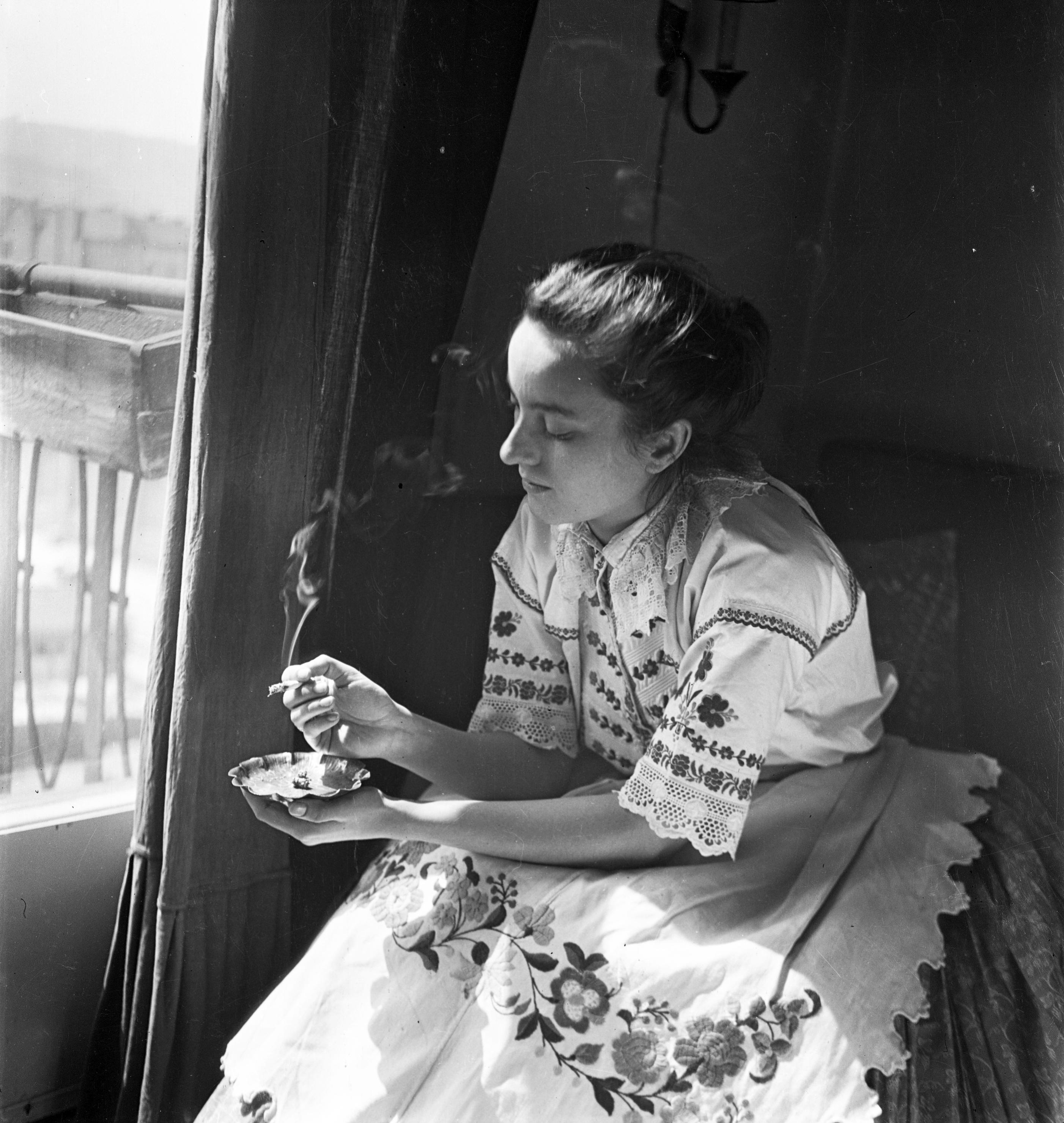 A young woman, dressed in traditional Hungarian folk costume, is sitting on a chair by  a window, smoking a cigarette in one hand, while the other hand holds an ashtray.  She has a serious, thoughtful look on her face as gazing downwards, rather than out the window.