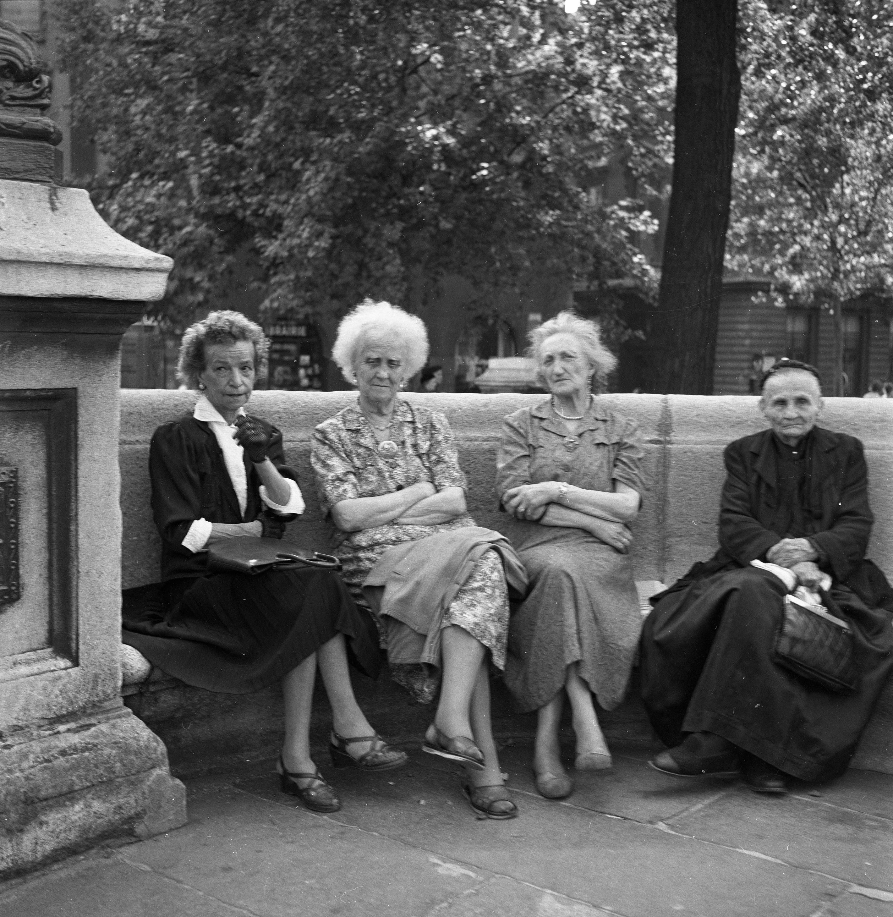 A black and white photo of four older women sitting on a curved stone bench in an urban setting.  The women look rather stern and unsmiling as if they have seen much suffering and heartache. One is rather elegantly dressed with gloves and has a satchel on her lap, while two others are in summer frocks, arms crossed. The fourth is dressed in all black suggesting she is a widow.