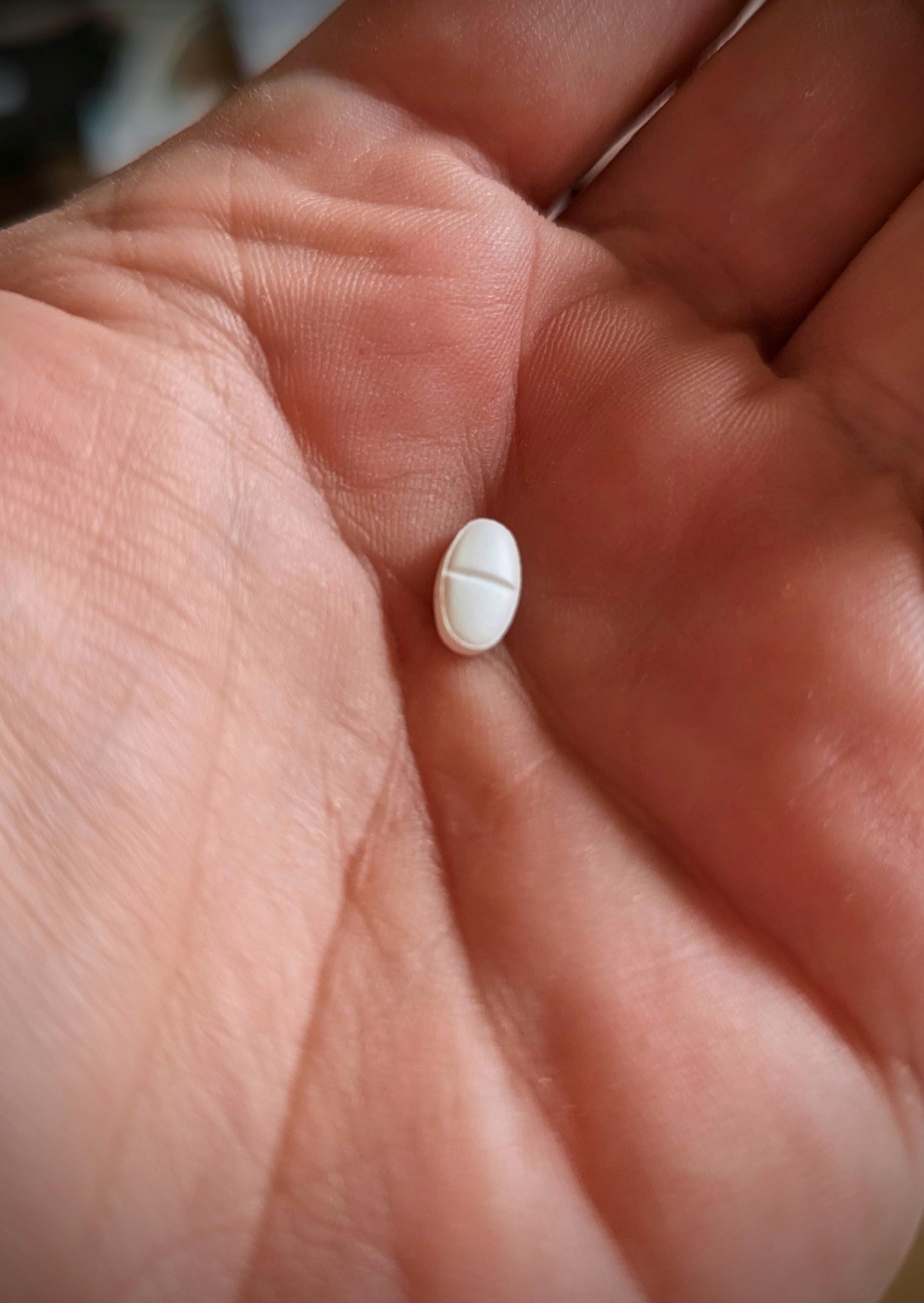 A close-up photograph of an open human hand holding a single, small, white oval pill with a score line down the middle resting in the palm.