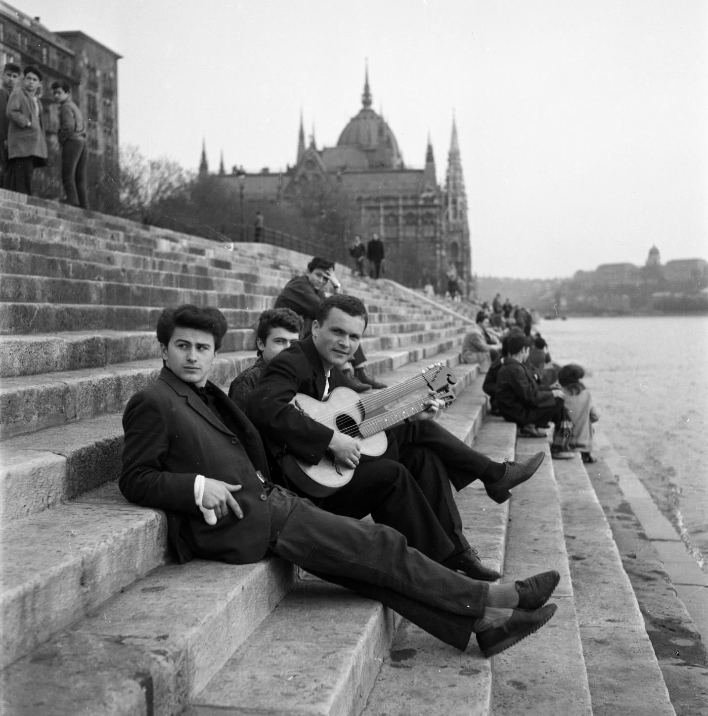A black and white photo of a group of young people sitting on the stepped embarkment of the river Danube.  One of the young men has a double-necked guitar on his knee. In the background are the Parliament building and further in the distance on the other side of the rive, the former royal castle can be seen.