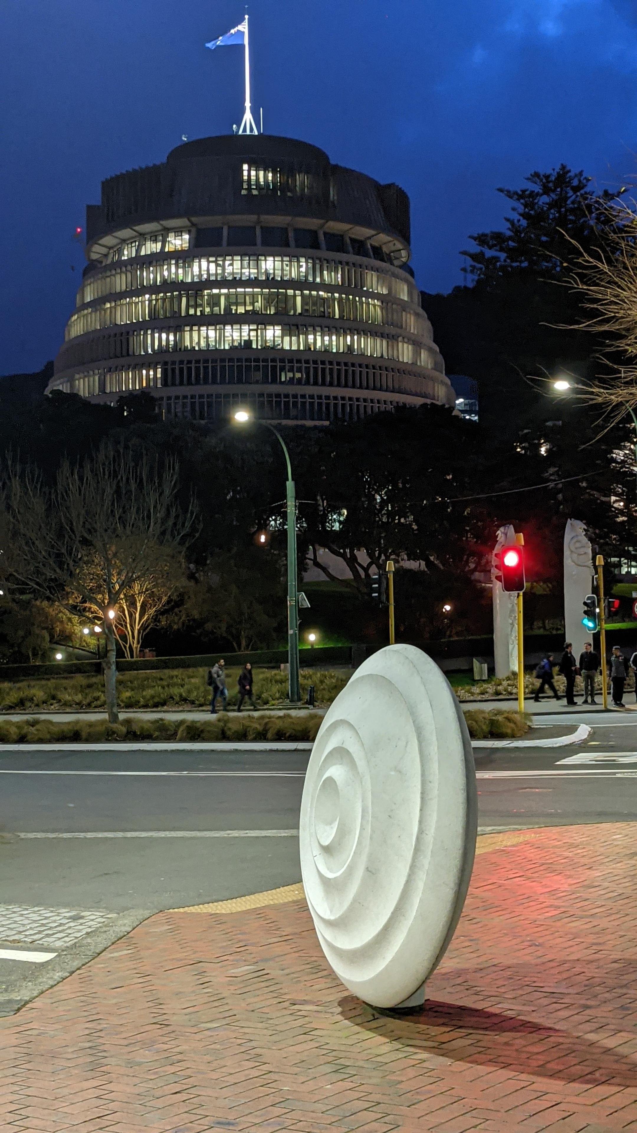 A vertical, slightly low-angle photograph of the Beehive, the Executive Wing of the New Zealand Parliament Buildings, at twilight, in Wellington, New Zealand. The unique, beehive-shaped, circular building is illuminated from within, with warm yellow light pouring from countless small rectangular windows across its multiple, diminishing-width floors, contrasting with the dark blue, cloudy evening sky. The building is made of concrete and glass. On the flat roof, the New Zealand flag, a blue banner with the Union Jack in the top left and four red-and-white stars on the right, is flying from a flagstaff. The building sits behind a dense layer of dark green trees, their leaves barely visible. In the midground, a paved street and pedestrian crossing are visible, framed by low, dark yellow-green shrubbery. Two pedestrians are walking along a path behind the shrubs. On the right, a streetlight and a traffic light on a tall pole are glowing green. In the foreground, positioned in the right of the frame on a red-brick sidewalk with red brick paving, stands a striking, contemporary white sculpture—a large, polished stone disk with deep, concentric spiral ridges carved into its surface. It leans slightly. In the far right, partially obscured, is another smaller white, shell-like sculpture. The mood is calm and evening-like. The focus is sharp on the foreground sculpture and the midground building. The overall lighting is twilight, mixing cool ambient light with warm artificial light.