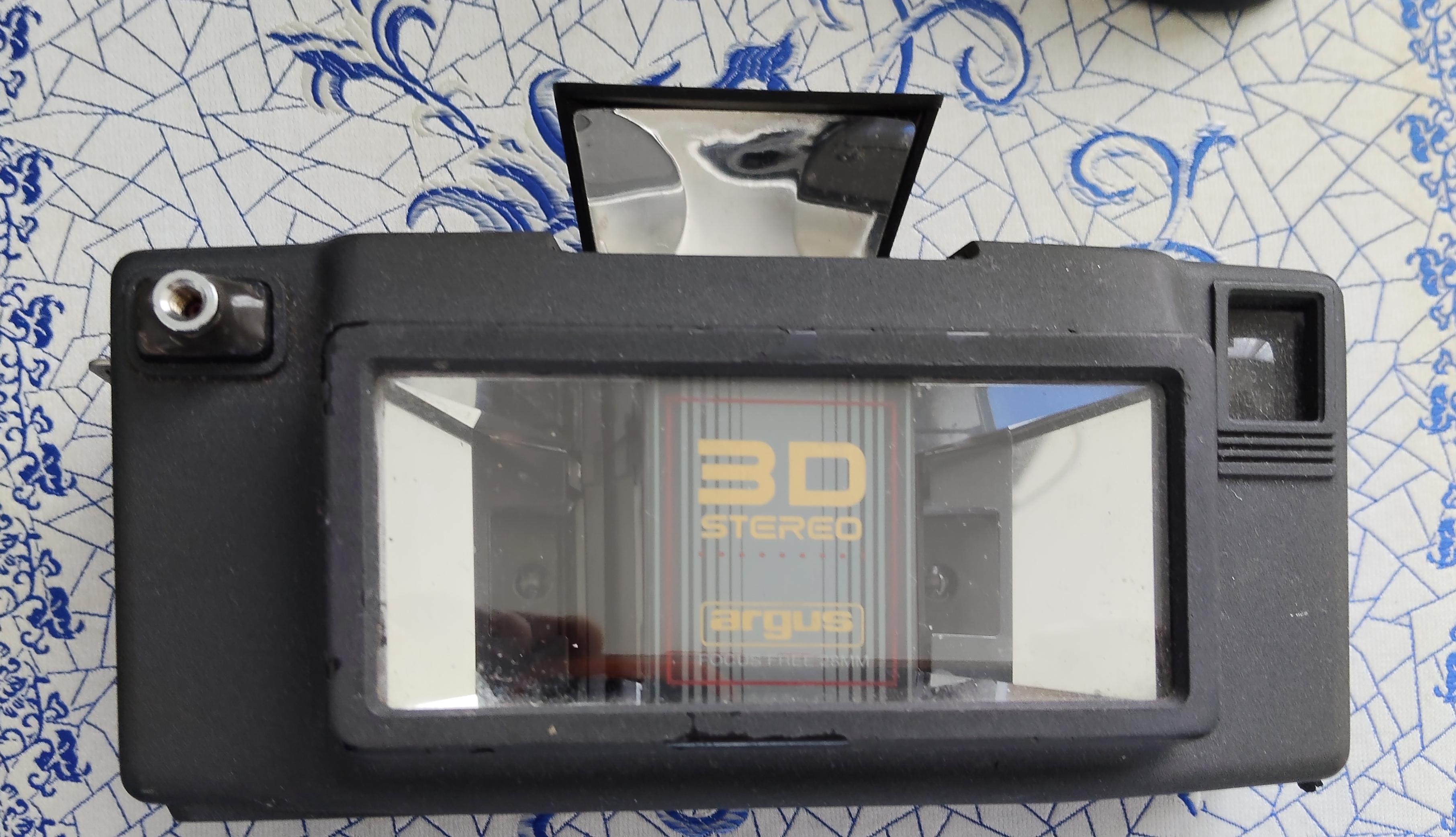 A top-down view of a rectangular, black matte plastic Argus 3D Stereo camera resting on a patterned cloth featuring blue mosaic and floral motifs.
Key features of the camera:
Centre: A large glass panel set into a raised rectangular frame. Behind the glass, gold lettering reads "3D STEREO" above a yellow "argus" logo, enclosed within a fine red and grey border. Internal mirrors inside the frame create visible reflections.
Top: A small, rectangular flash unit popped up from the top edge of the camera body, showing a shiny metallic reflector inside.
Top Left: A small metal threaded shutter release button on a raised mound.
Top Right: A small rectangular glass viewfinder set into the camera body.
