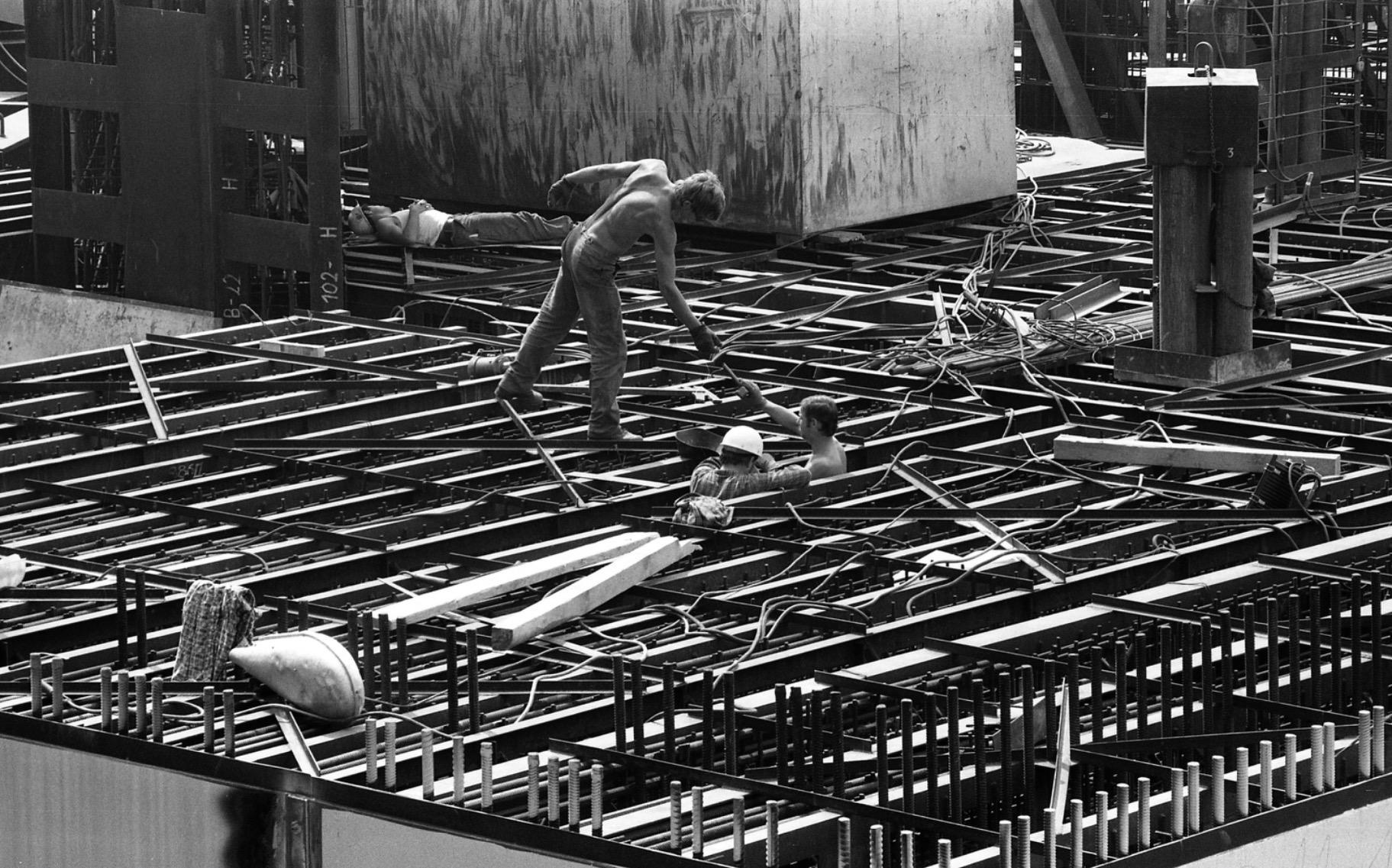 A black and white photo of a large building site show several workers, including one who appears to be lying down napping, working on the reinforcing steel and steel girders.
