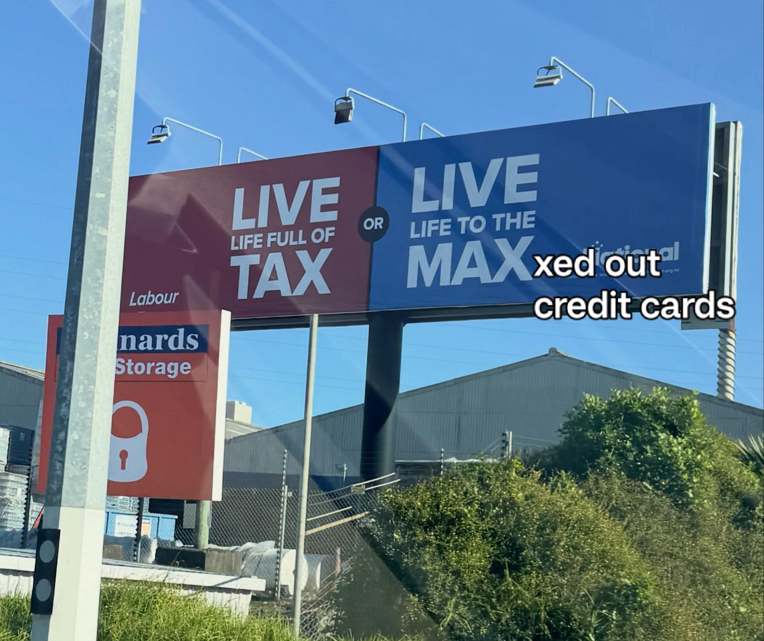 A roadside billboard divided into two halves. The left side is red with white text reading "LIVE LIFE FULL OF TAX" above the Labour Party logo. The right side is blue with white text reading "LIVE LIFE TO THE MAX" above the National Party logo, but white text with a black outline overlaying the lower right corner modifies it to read "xed out credit cards" so the full blue section reads "LIVE LIFE TO THE MAXed out credit cards".