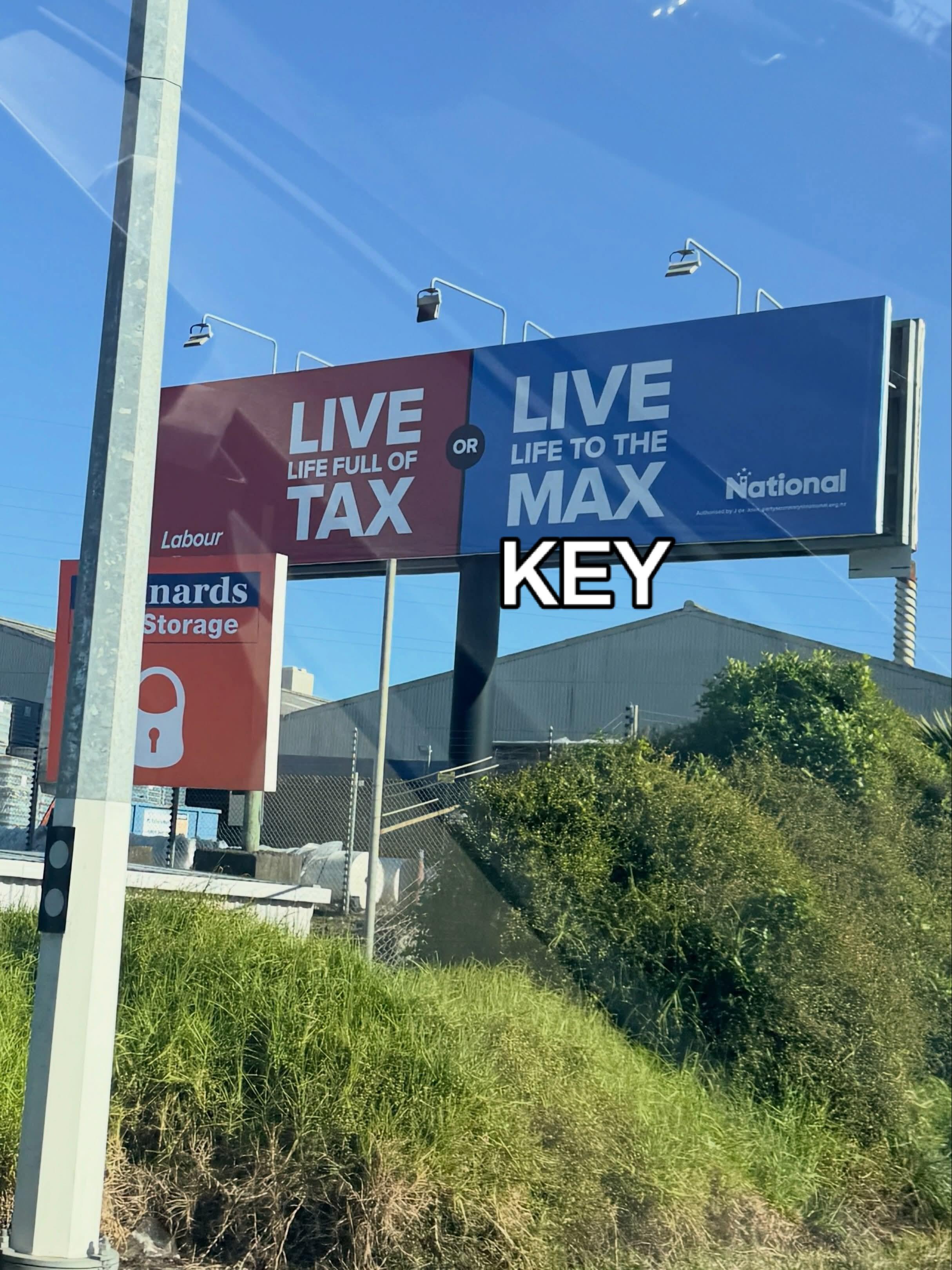 A roadside billboard split into two halves. The left side is red with white text reading "LIVE LIFE FULL OF TAX" above the Labour Party logo. The right side is blue with white text reading "LIVE LIFE TO THE MAX" above the National Party logo, with white text outlined in black overlaying the bottom right corner that reads "KEY", modifying the blue section to read "LIVE LIFE TO THE MAX KEY".