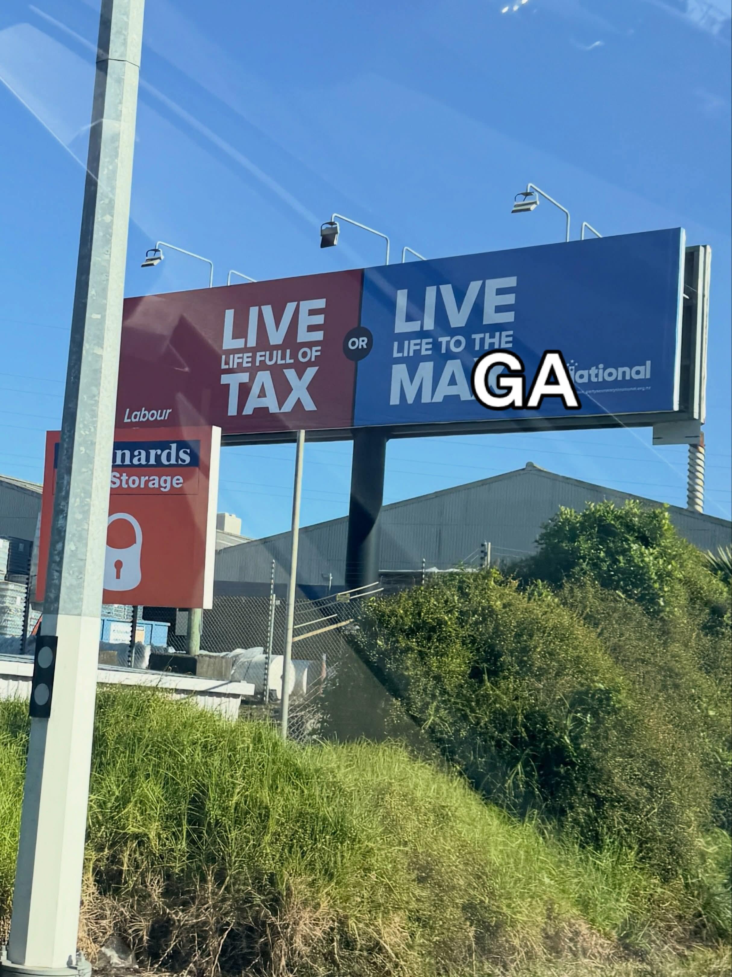 A roadside billboard divided into two halves. The left side is red with white text reading "LIVE LIFE FULL OF TAX" above the Labour Party logo. The right side is blue with white text originally reading "LIVE LIFE TO THE MAX" next to the National Party logo, but white text with a black outline has been overlaid to replace the "X" with "GA", modifying the full blue section to read "LIVE LIFE TO THE MAGA".