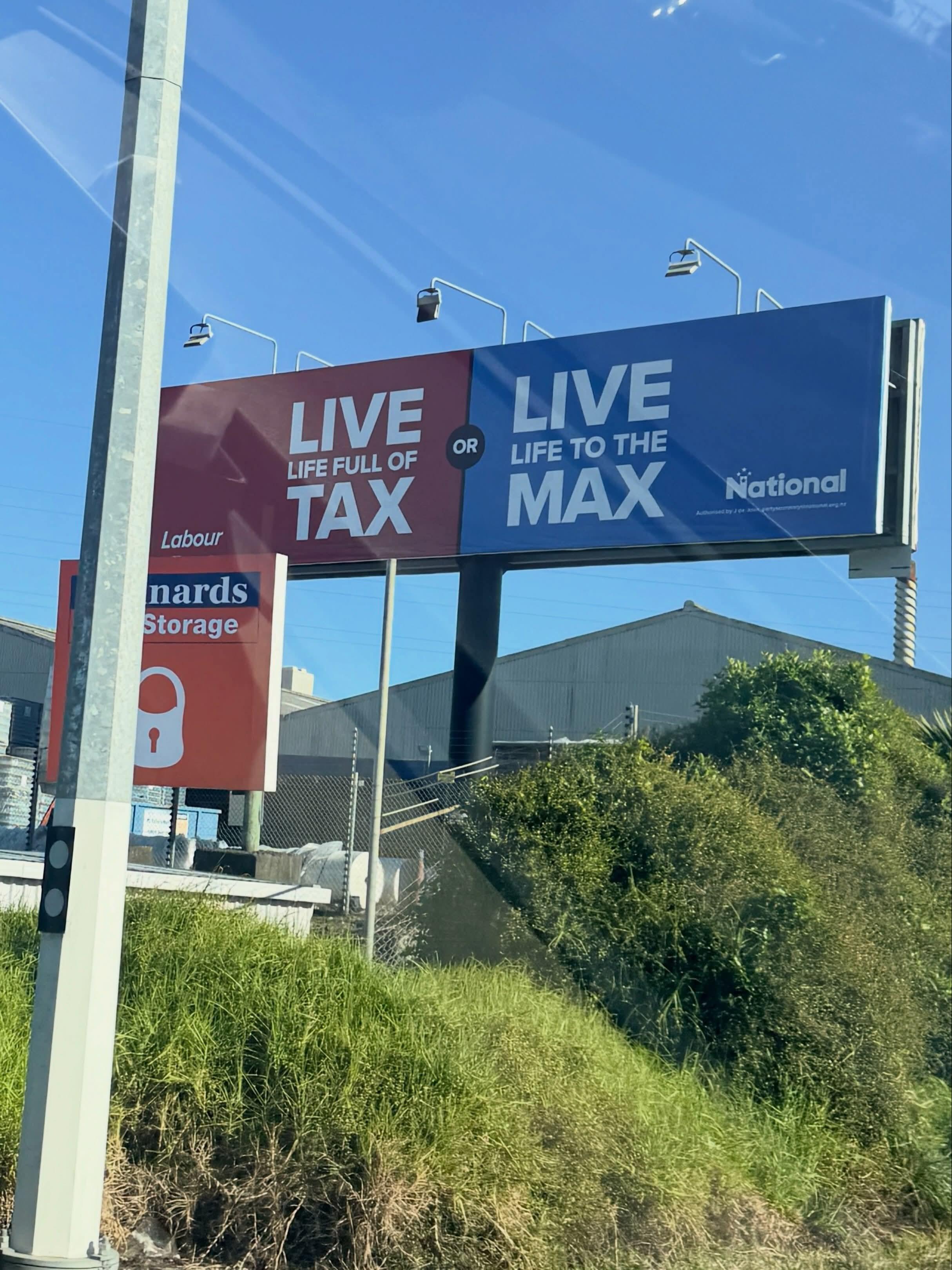A political billboard split into two contrasting halves against a clear blue sky. The left half is red with large white text that reads "LIVE LIFE FULL OF TAX" above a small Labour Party logo. The right half is blue with large white text reading "LIVE LIFE TO THE MAX" next to a white National Party logo. A small black circle in the centre separating the two sides contains the word "OR".