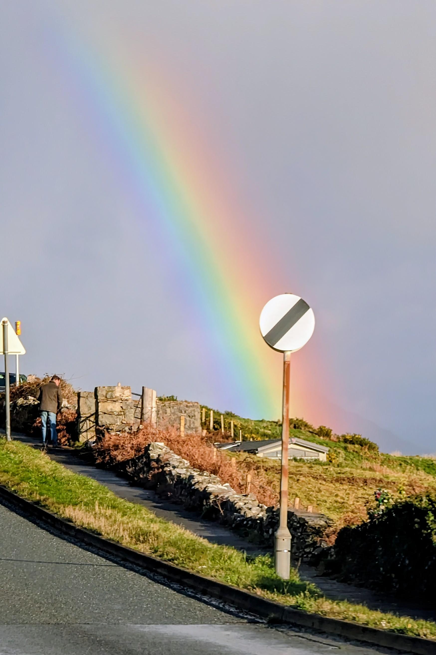 A photograph looking towards rolling hills from the Porthmadog Road in Criccieth, North Wales, under a moody gray sky after a shower. A vibrant, well-defined rainbow spans the sky. In the foreground, on the right, is a circular white UK road sign with a diagonal black stripe. On the steep uphill sidewalk to the left, a man in a dark jacket and jeans walks past a low, weathered dry-stone wall and a gorse-covered bank. A few distant houses from the town are visible on the hillside.