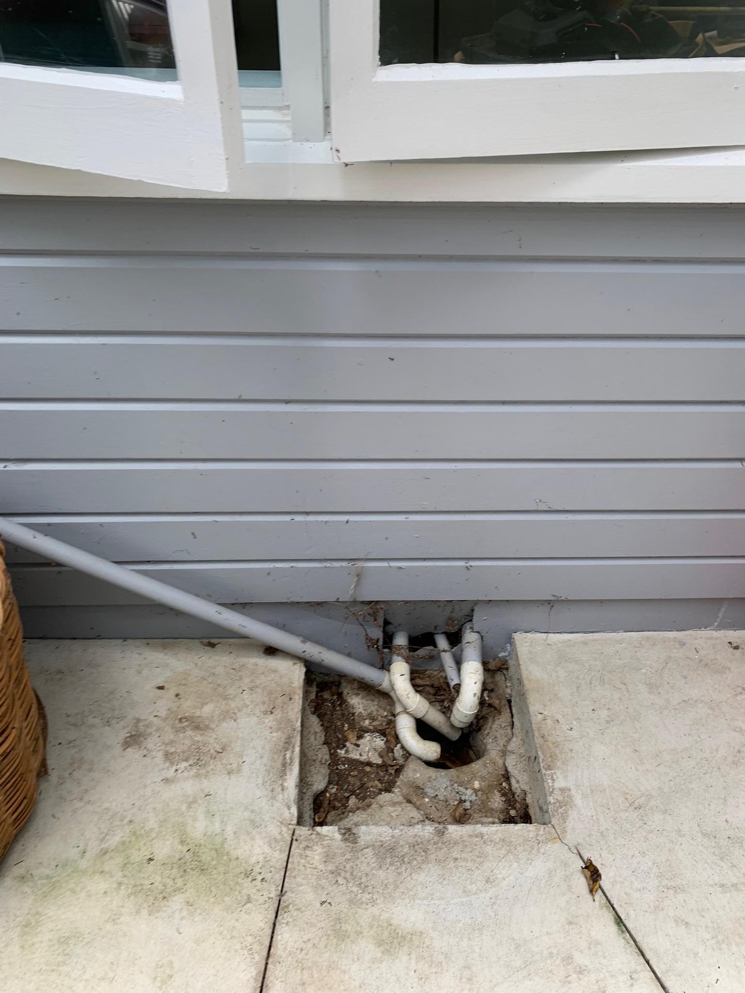 Various water pipes coming out from the side of a house and going in to a hole in the ground. There’s a big square of space around them before the smooth concrete starts.